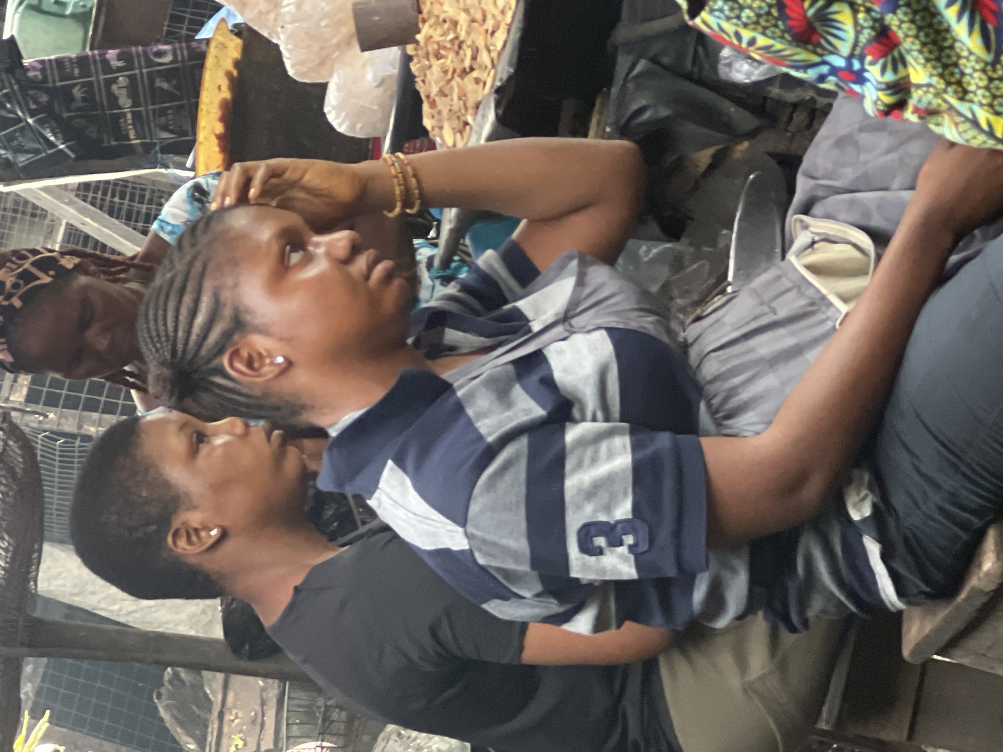 The image captures a scene at a market in Onitsha, Nigeria. In the foreground, a woman with braided hair is seated, wearing a blue and gray striped shirt with the number "3" visible on the sleeve and a gray garment. She has bracelets on her wrist and is touching her forehead. To her left, another person wearing a black t-shirt is partially visible. In the background, another woman with a patterned headscarf is seen, along with market stalls displaying various goods, including what appears to be dried food items. The environment suggests an outdoor or semi-covered market setting.
