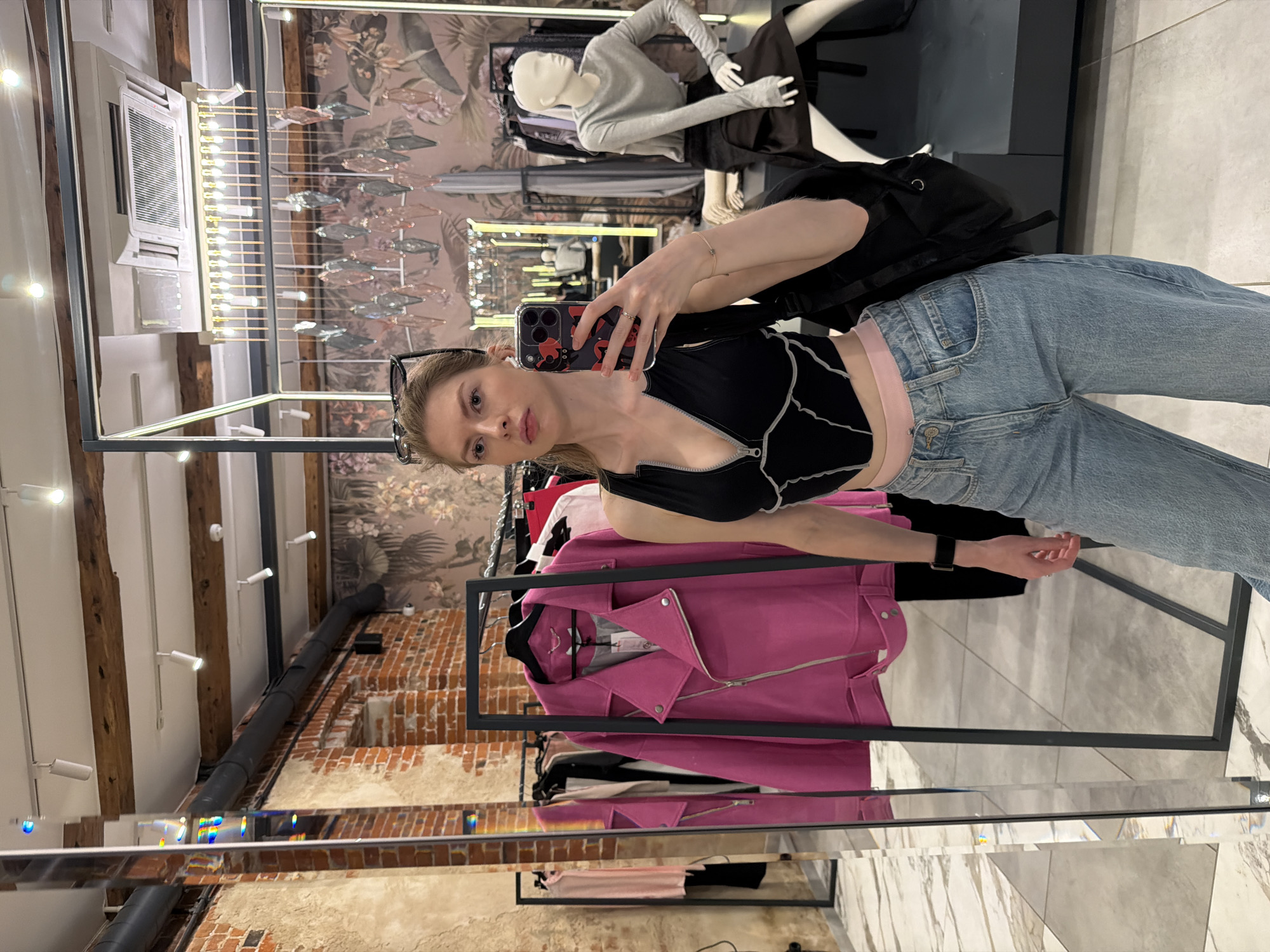 A young woman is taking a selfie in what appears to be a clothing store. She's wearing a black top with a unique stitch design, light-wash jeans, and has a black backpack slung over her shoulder. Her sunglasses rest on her head.
Behind her, racks of clothes are visible, including a prominent pink jacket. A mannequin is partially visible in the background. The architecture of the building is visible.
Given the architectural details, it could be in a historical part of Moscow, Russia. Without more specific details, it is impossible to pinpoint the exact location in the city.
