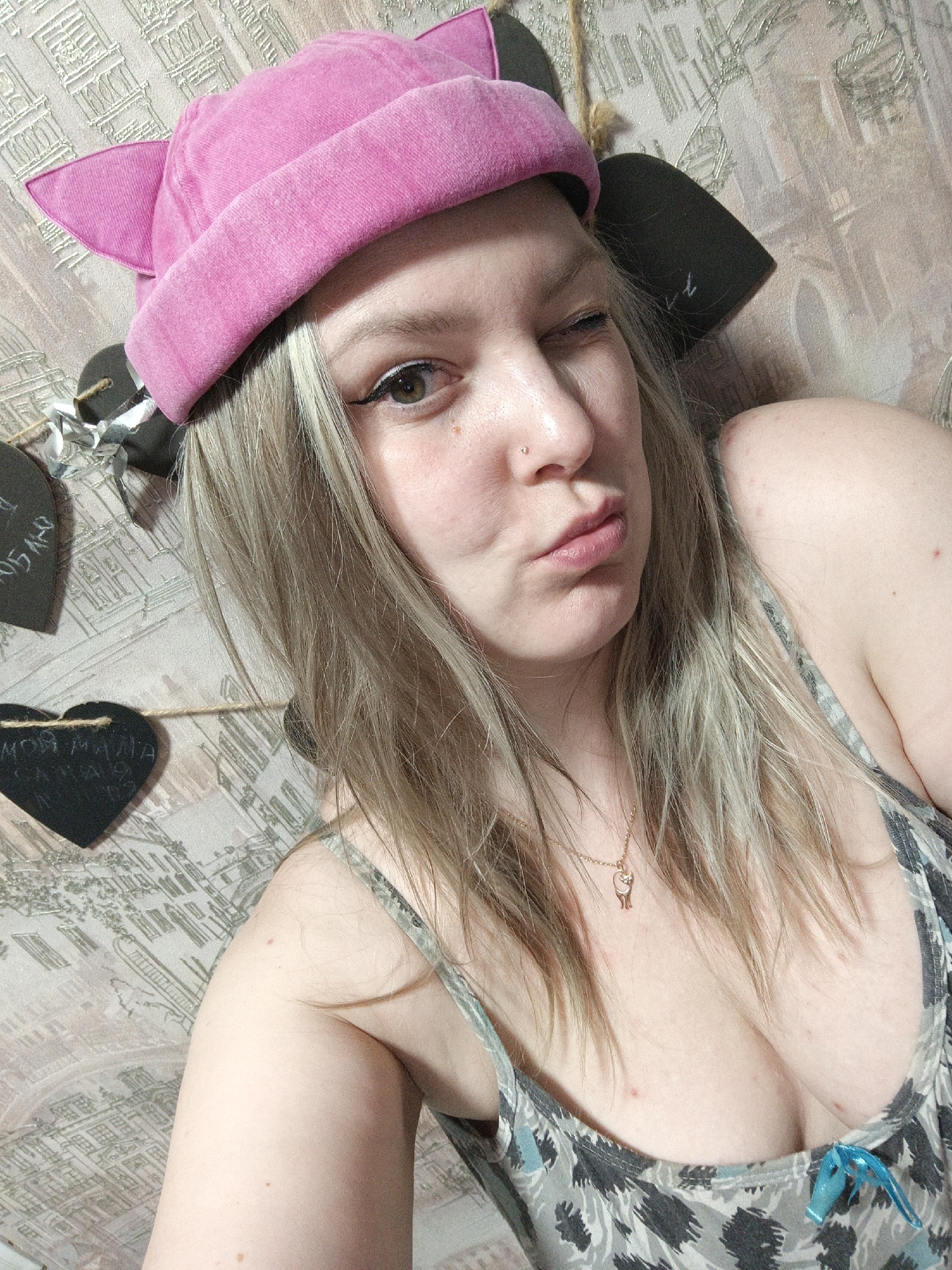 The media file presents a close-up selfie of a young woman in what appears to be a casual indoor setting, located in Марковский, Russia. She has long, light blonde hair with darker roots, which falls over her shoulders and chest. She is wearing a bright pink hat shaped like cat ears and a patterned tank top or camisole, which features a blue bow detail near the cleavage. A delicate gold necklace with a small pendant hangs around her neck.
The woman is captured mid-expression, winking with her right eye and pouting or puckering her lips. A small stud piercing is visible on her left nostril. Her right eye shows visible black eyeliner. Her complexion is fair, with some natural skin texture and a few small red marks or blemishes visible on her chest and shoulder.
The background is a wall covered with light-colored wallpaper featuring a repeating architectural pattern, possibly depicting buildings or facades. Several decorative heart-shaped objects are strung along a twine or string across the wall. Two of these hearts are black and appear to be chalkboard material. Visible Russian text on one black heart reads "МОЯ МАМА САМАЯ" (My mom is the most...), with the rest cut off. Another partial inscription above it seems to be "Я ЛЮБЛЮ" (I love...). One of the hearts also has silver tinsel or foil attached. The lighting is bright and even, indicating an indoor environment. The overall mood conveyed is playful and casual.