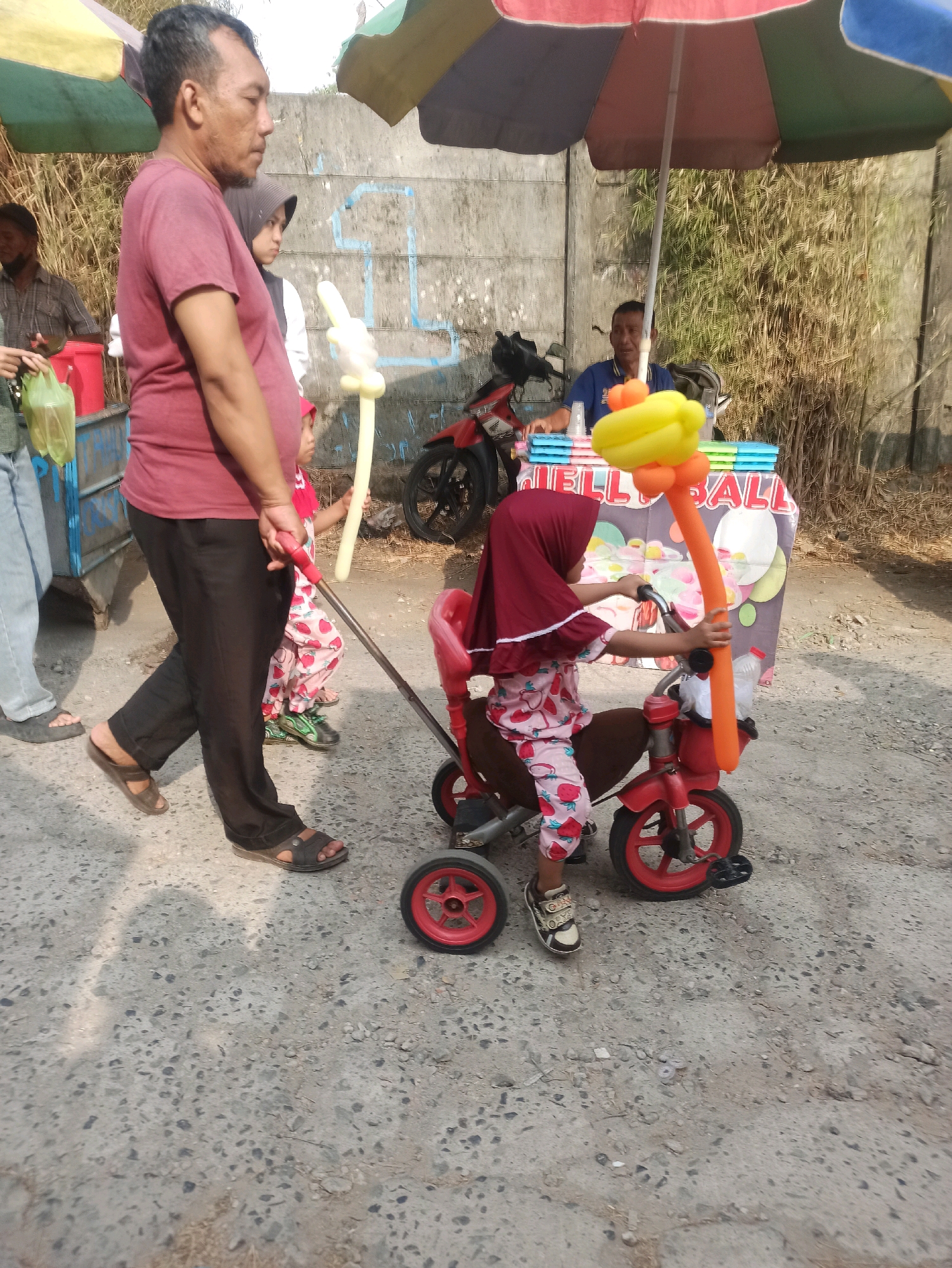 A man, wearing a maroon t-shirt and black pants, walks on a paved road next to a food stall. Behind him, a young girl in a pink and white strawberry-patterned outfit and a maroon hijab rides a red tricycle. The tricycle has balloon decorations. A vendor is visible behind the stall, which has colorful signage. A motorcycle is parked near the stall. The background shows a concrete wall and dry vegetation.