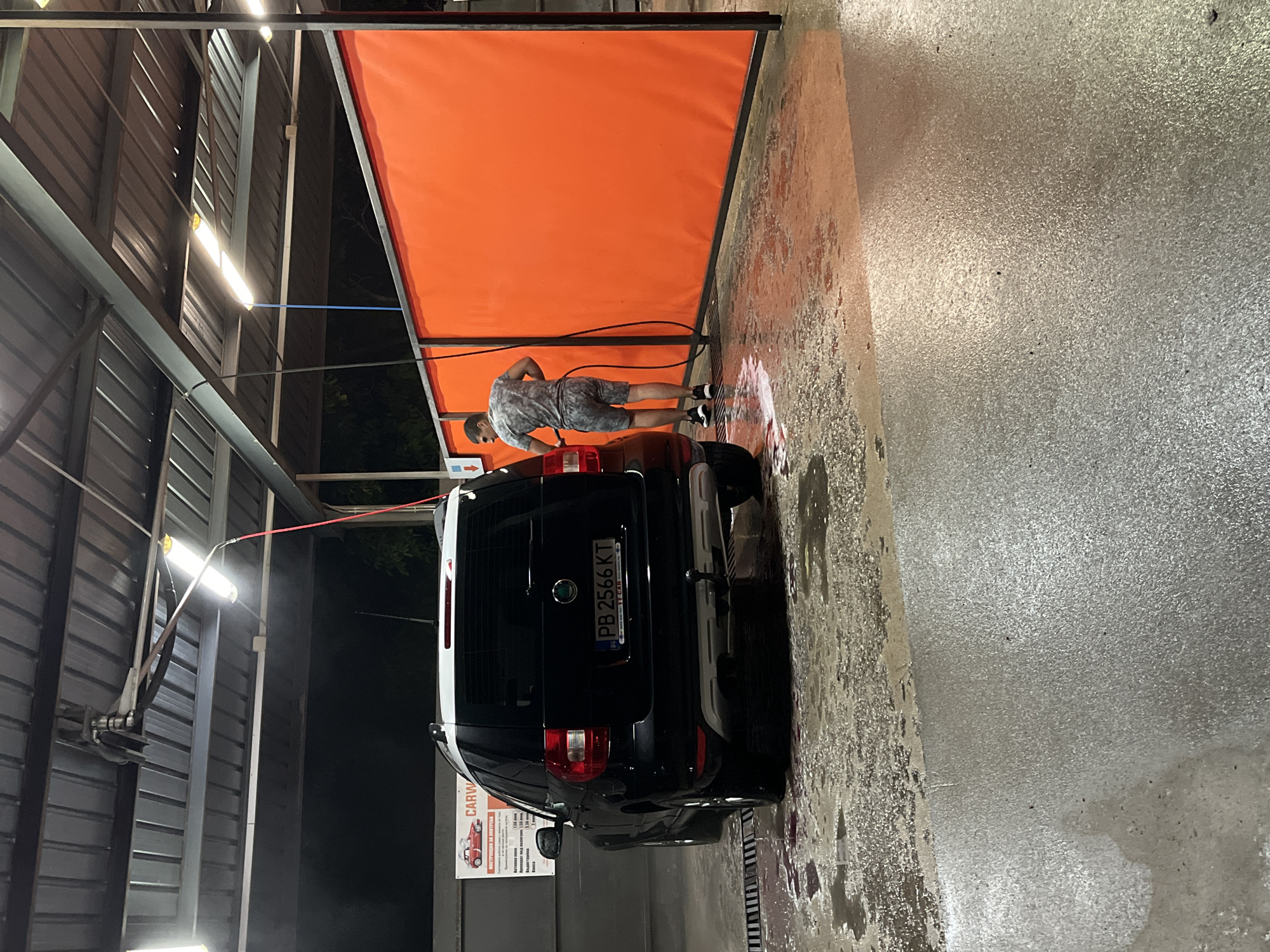 The image depicts a self-service car wash facility at night, likely located in Anevo, Bulgaria. In the foreground, a black Skoda Yeti is parked, with its rear facing the viewer. The car has a white roof and a license plate reading "PB 2566 KT." The ground is wet and reflects the overhead lighting, indicating recent or ongoing activity. To the right of the car, a man wearing a camouflage-patterned t-shirt and shorts is using a high-pressure hose to wash the vehicle. He is positioned between the car and a large, orange fabric barrier that separates the car wash bays. Overhead, a metal roof structure with fluorescent lights provides illumination. A sign is visible on the left wall, partially obscured, showing graphics related to car washing services. The overall environment suggests a functional, utilitarian space.
