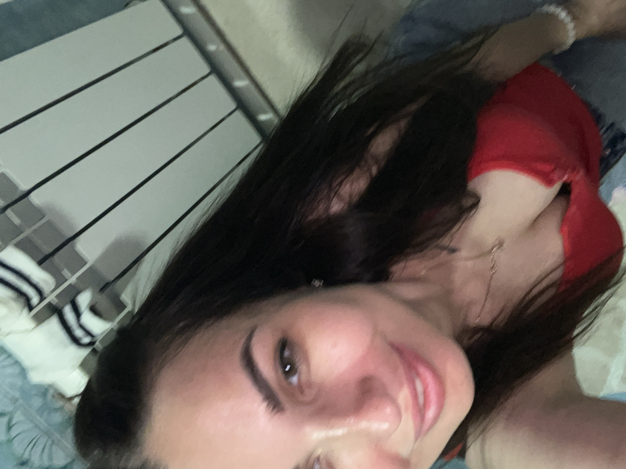 A woman with long, dark hair is taking a selfie. She is wearing a red top and a necklace. Her hair is partially covering her face, and she is smiling. The background appears to be indoors, with some sort of white shelving unit with dark stripes visible.