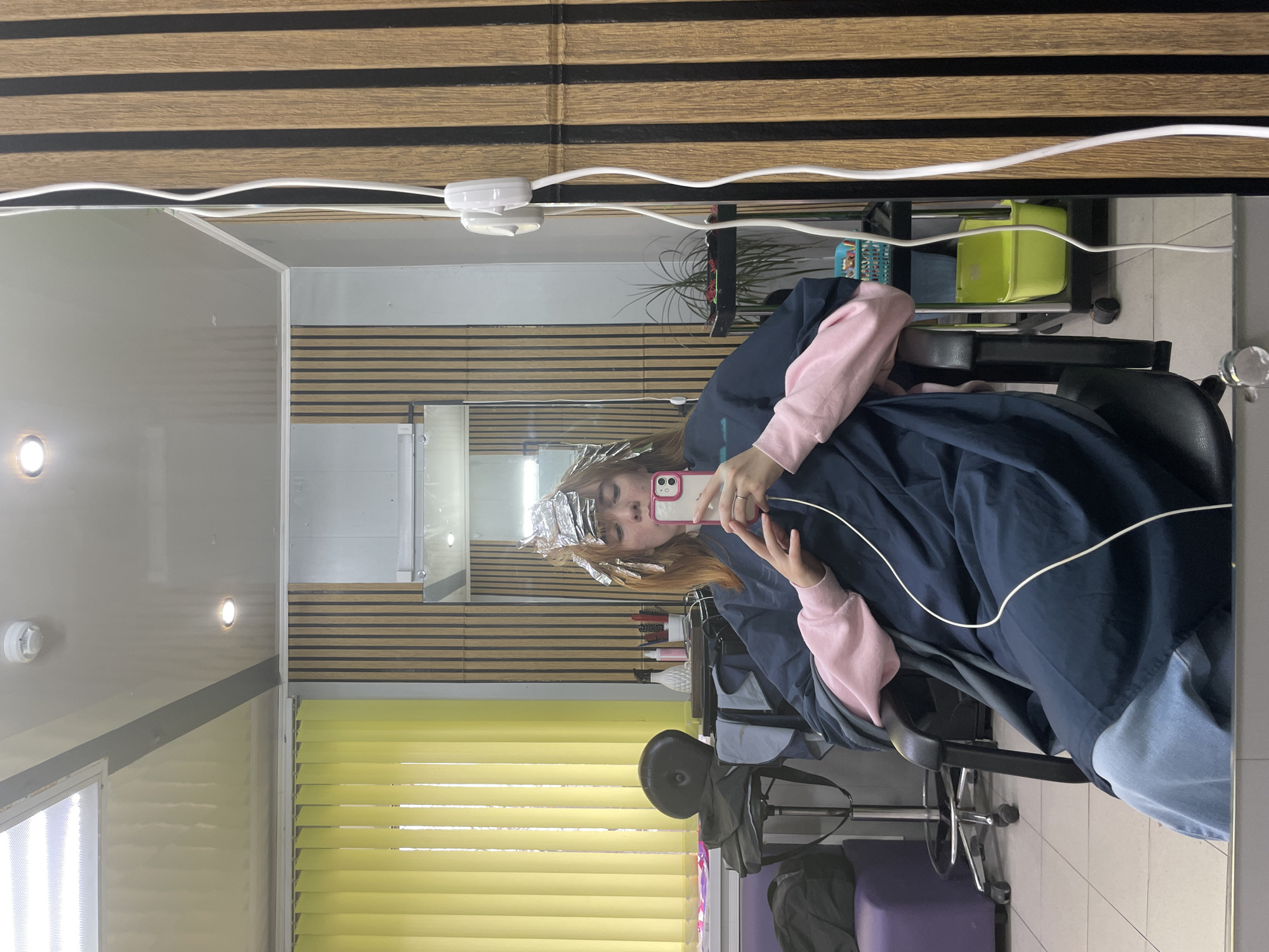 A mirror selfie captures a young person receiving a hair treatment in a salon in Chaikovsky, Russia. The image is oriented sideways, showcasing the reflection of the person and the salon interior.
The subject, a young individual, is seated in a black salon chair with wheels, taking a photo of their reflection with a smartphone that has a white and pink case. Their hair is partially covered with metallic foil, indicating a highlighting or coloring process. They are dressed in a dark blue protective smock over a pink long-sleeved top and light blue jeans.
The salon environment, visible in the mirror's reflection, features a wall decorated with wooden slat paneling. A window with bright yellow horizontal blinds is visible, suggesting natural light. Various salon items are present, including a metal shelving unit holding bottles and brushes, a green plastic container, and a potted plant. A white electrical cable snakes across the upper portion of the reflected image. The ceiling contains recessed lights, and a white smoke detector is mounted on a wall. The floor is tiled with light-colored squares. The scene depicts a modern hair salon where a client is undergoing a beauty treatment.