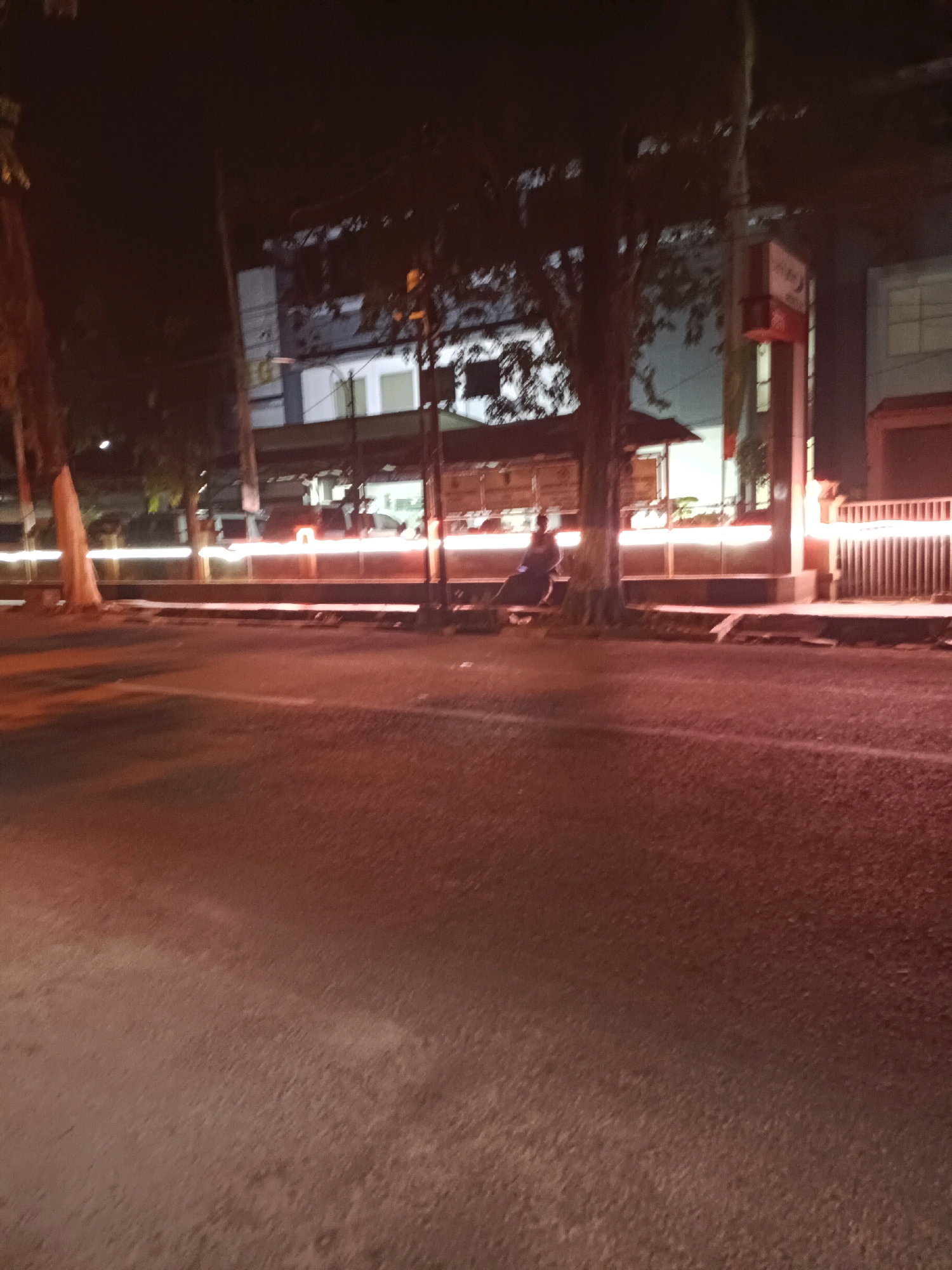 This image depicts a street scene at night, likely in Lamongan, Indonesia, given the context provided. The foreground is dominated by a dark, asphalt road, with some blurring suggesting motion or low light conditions. In the mid-ground, there's a sidewalk area with a tree and a person seated on a bench or the ground under it. Beyond the sidewalk, a building with illuminated windows and signage is visible. A fence with vertical bars and lights runs along the right side of the mid-ground. Horizontal streaks of light, possibly from passing vehicles, are visible across the scene. The overall atmosphere is dark and urban.