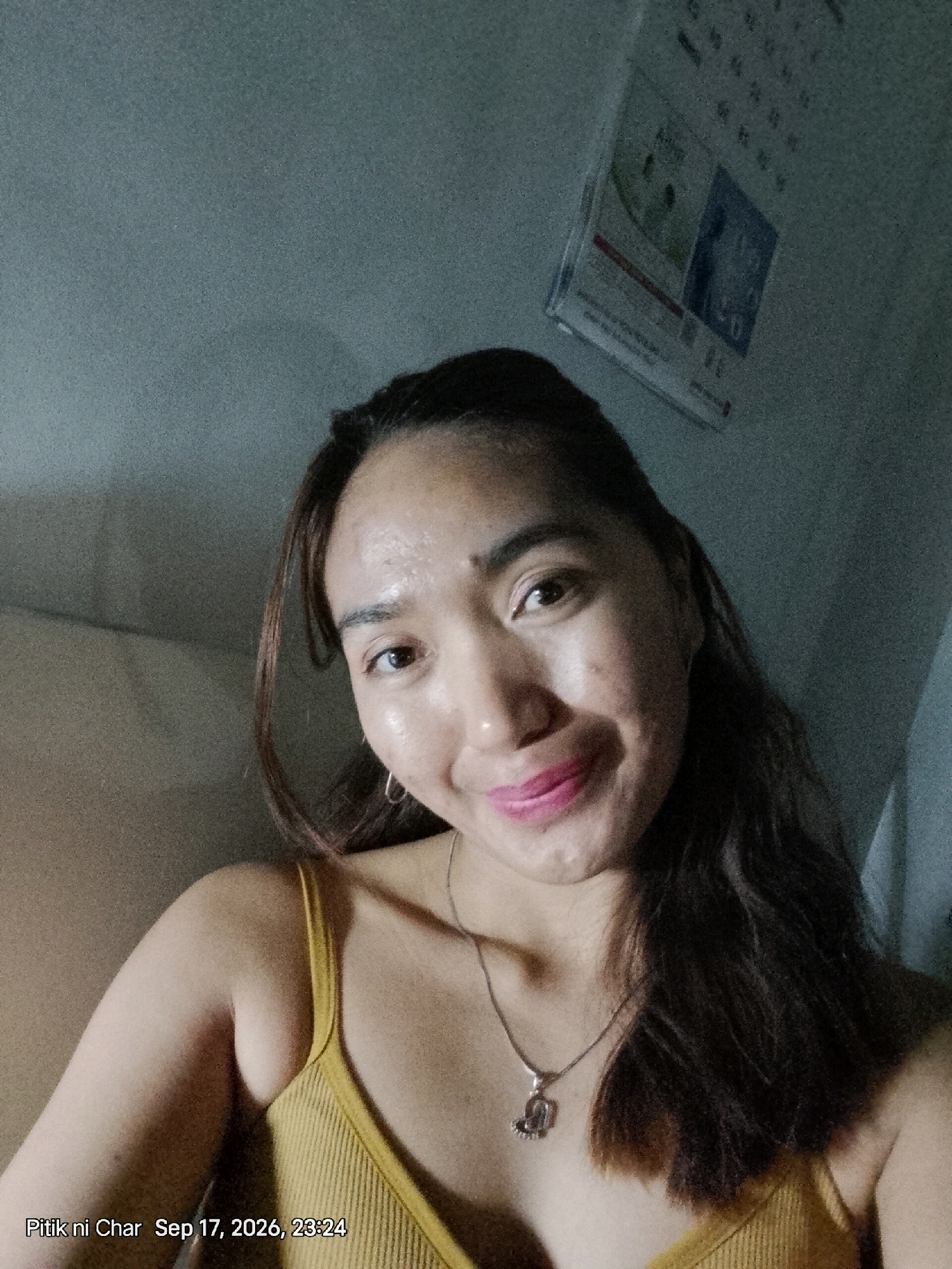 A woman with dark hair is taking a selfie in a room. She is wearing a yellow ribbed tank top and a silver necklace. She has on pink lipstick and is smiling. The background is a gray wall with a calendar hanging on it. The date on the calendar appears to be September 17, 2026, 23:24. The location is identified as Davao City, Philippines.