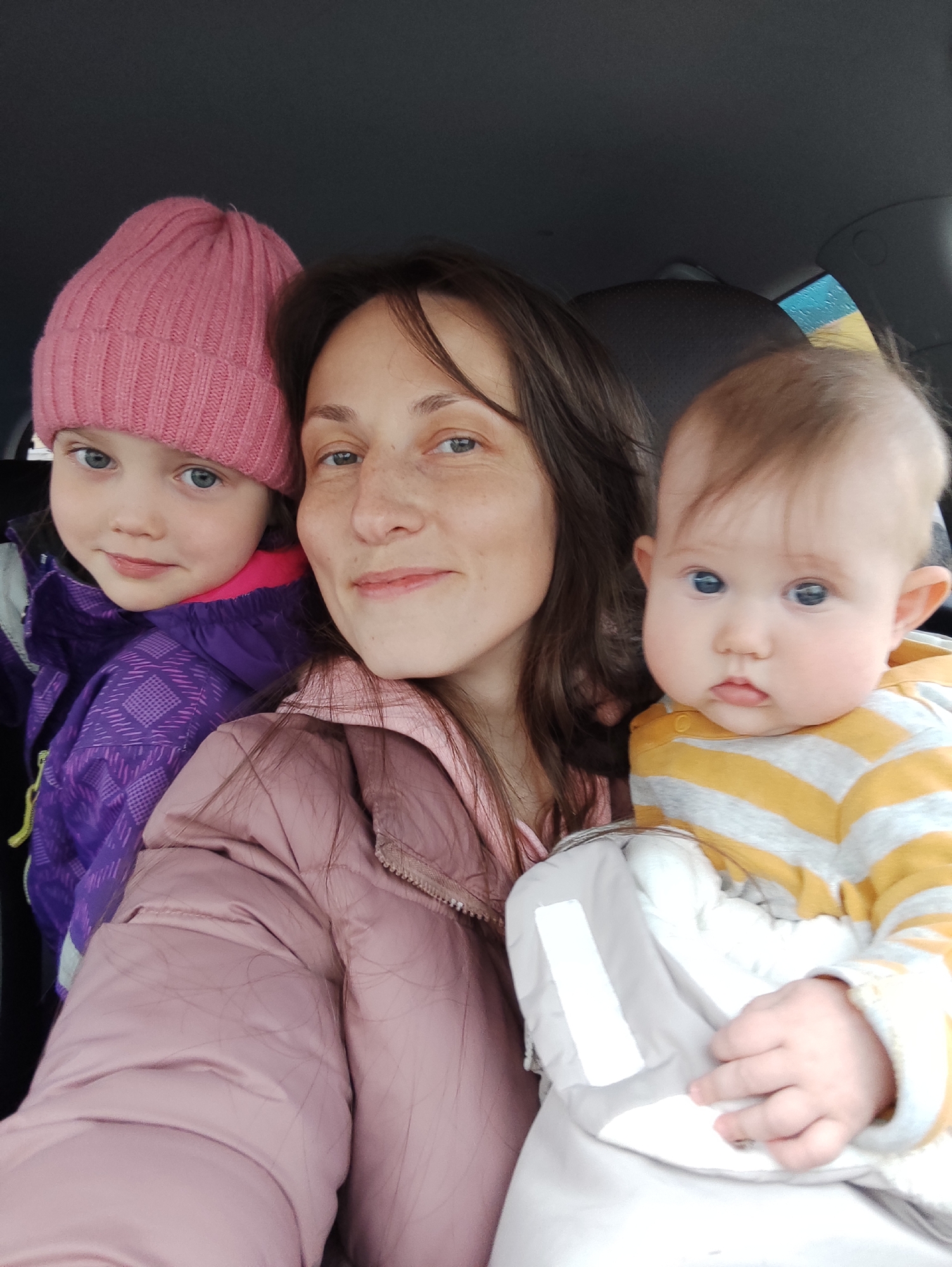 A selfie shows a woman with two young children in a car. The woman, in a pink jacket, is centered in the frame. To her left is a girl wearing a pink hat and purple jacket. To the woman's right is a baby in a yellow and gray striped shirt, wrapped in a gray blanket. The backdrop is the interior of a car. The location is likely Chaikovsky, Russia, based on prior knowledge.