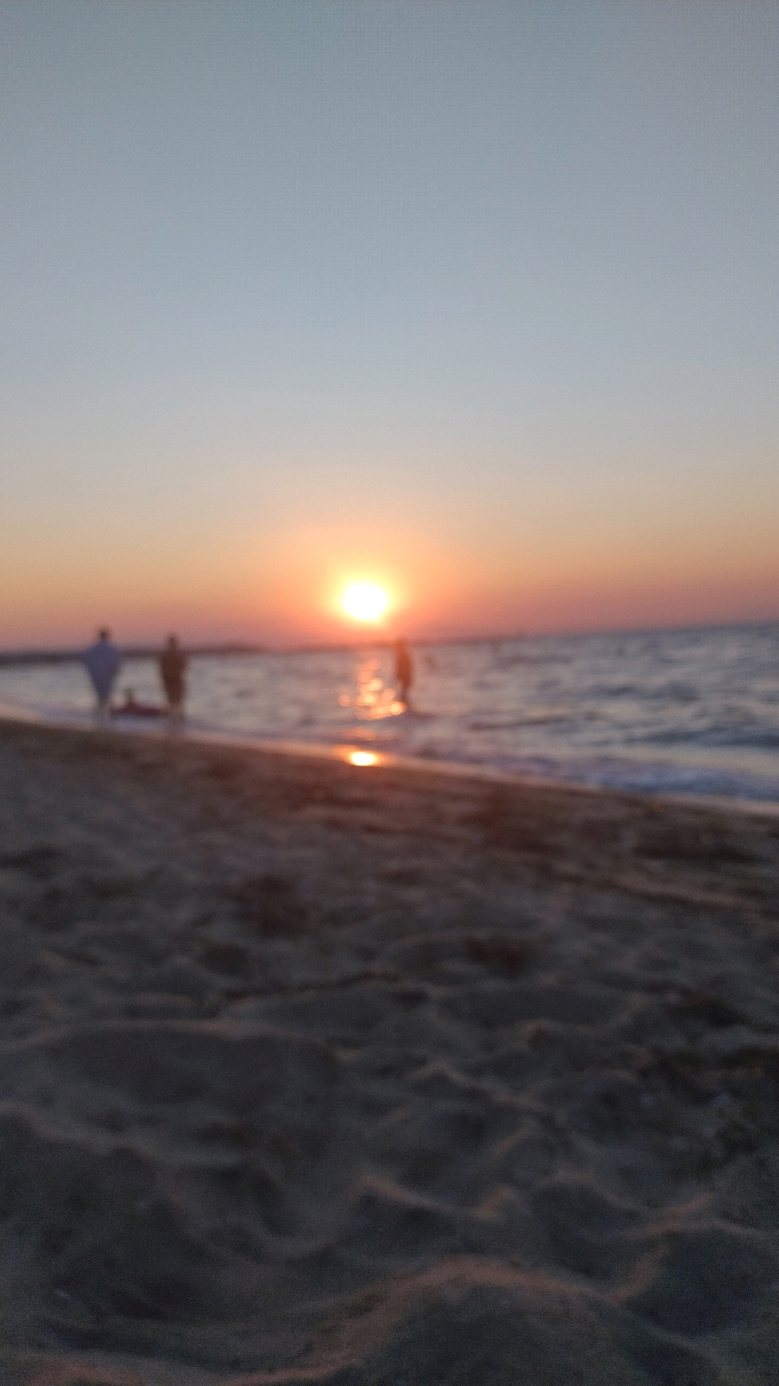 The image depicts a beach scene at sunset in Neoi Epivates, Greece. The foreground is a sandy beach, appearing out of focus. In the middle ground, a body of water, likely the sea, reflects the sunset. Several indistinct figures of people are visible on the shoreline and in the shallow water. The sun is low on the horizon, casting a warm, orange glow that illuminates the sky and water. The overall impression is of a tranquil evening at the coast.
