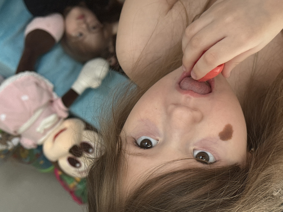 An overhead, slightly angled shot depicts a young female child with light brown hair, lying on her back. She looks directly at the camera with wide brown eyes. Her mouth is open, with her tongue extended, holding a small, red, oval-shaped object, possibly a piece of candy. Her right hand, partially visible, grips the object. A distinct, dark brown, heart-shaped mark is visible on her left cheekbone. Behind her head, an out-of-focus Minnie Mouse plush doll, wearing a pink dress, lies on its back. Further in the background, to the right of the main subject, a second, heavily blurred figure, appearing to be another child or doll, is visible lying down. The subjects are positioned on a light blue surface, likely bedding or a blanket, in an indoor setting.