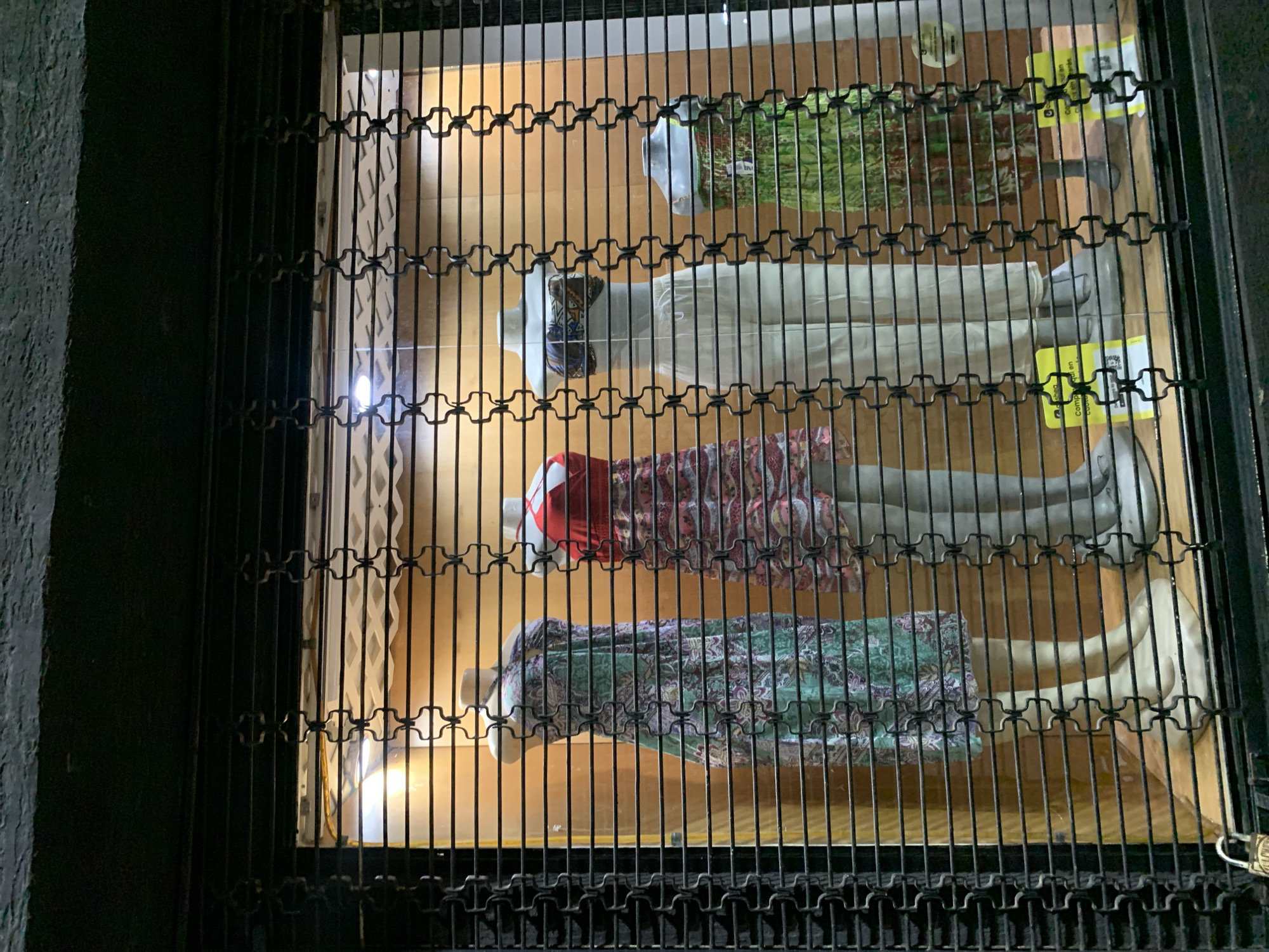 A window display protected by an ornate iron gate features four mannequins dressed in different outfits. The display is illuminated from within. The location appears to be in Juan Griego, Venezuela.