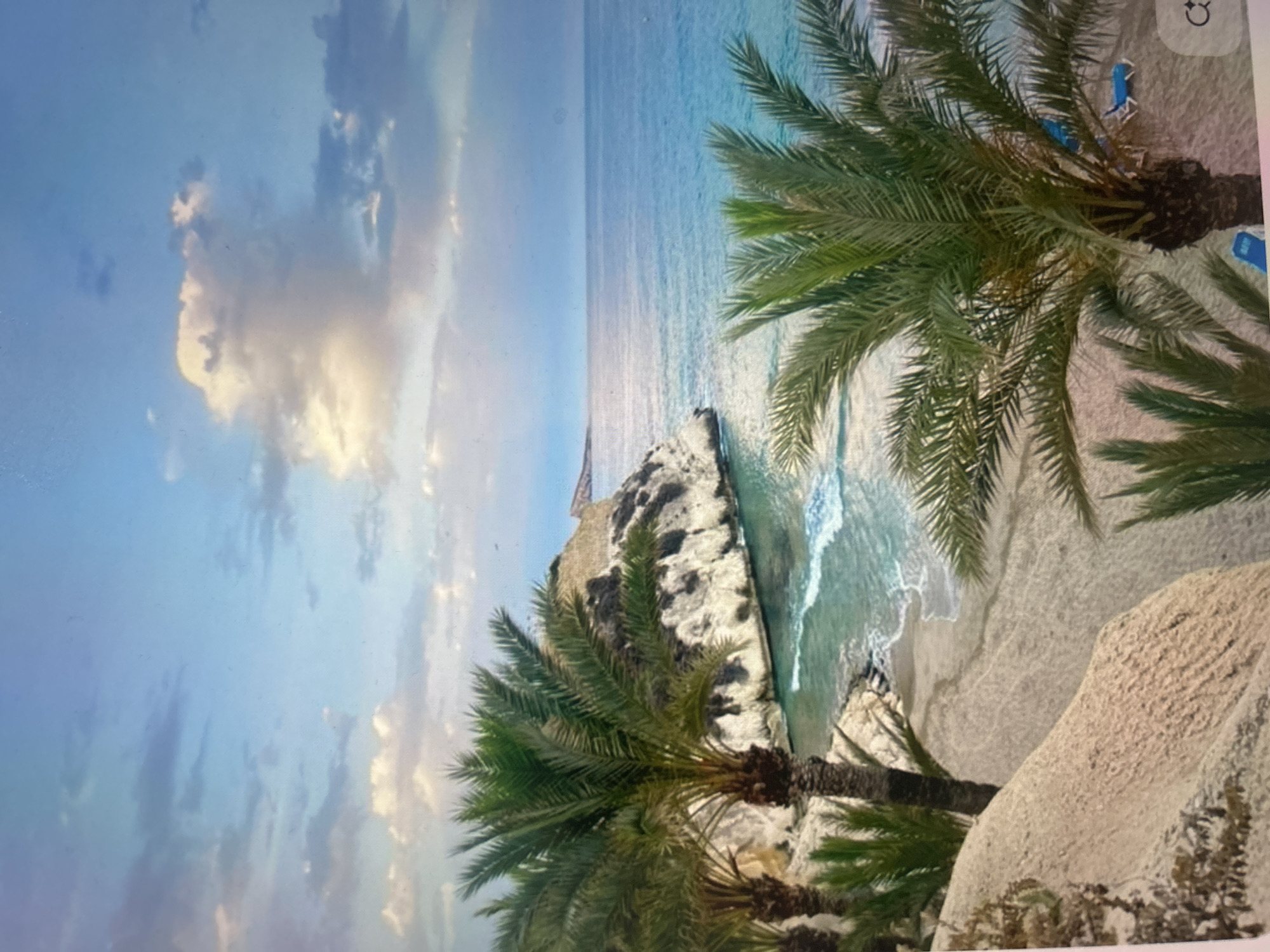 The image is a landscape photograph, displayed as if rotated 90 degrees clockwise, depicting a picturesque coastal scene.
If viewed in its intended horizontal orientation, the upper left quadrant of the frame is dominated by a bright blue sky, adorned with scattered, fluffy cumulus clouds. These clouds exhibit warm, golden-white highlights, suggesting the soft light of late afternoon or early evening.
The right side of the image features a calm, clear ocean, transitioning from turquoise near the shore to deeper blues further out towards the horizon. Gentle waves are seen breaking onto a light sandy beach. A prominent, light-colored rocky cliff or headland juts out into the water, with a small, weathered structure perched on its summit.
Several vibrant green palm trees are strategically placed, framing the scene. A large palm occupies the upper right portion (when viewed horizontally), with its fronds extending downwards. Below this palm, two blue beach chairs are subtly visible on the sand, indicating a recreational or leisure setting. Another palm tree is partially visible in the bottom center. The foreground on the lower right consists of light-colored sand and rocks.
The overall atmosphere conveyed is one of serene tropical or Mediterranean beauty, characterized by clear skies, tranquil waters, and lush coastal vegetation. A small icon, possibly related to image viewing controls (resembling a refresh or rotate symbol), is faintly visible in the top right corner of the original image capture interface.