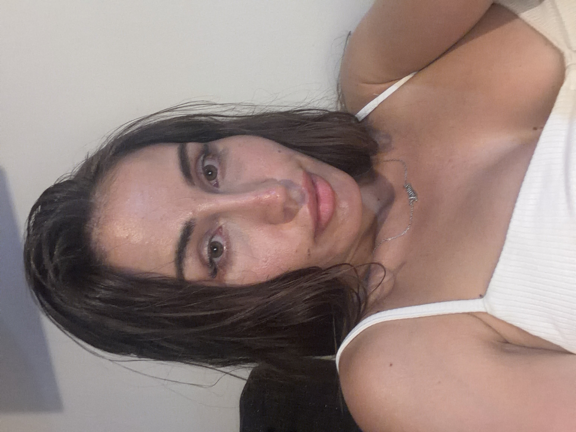 The image is a close-up selfie of a woman with dark brown hair and brown eyes. She is wearing a white tank top and a silver necklace. The background appears to be an indoor setting with a plain wall. The lighting suggests the photo was taken indoors, possibly at night. The woman's face is partially illuminated, casting shadows. There are no identifiable landmarks or features that would indicate a specific location within Tersefanou, Cyprus. The focus is solely on the woman's upper body and face.