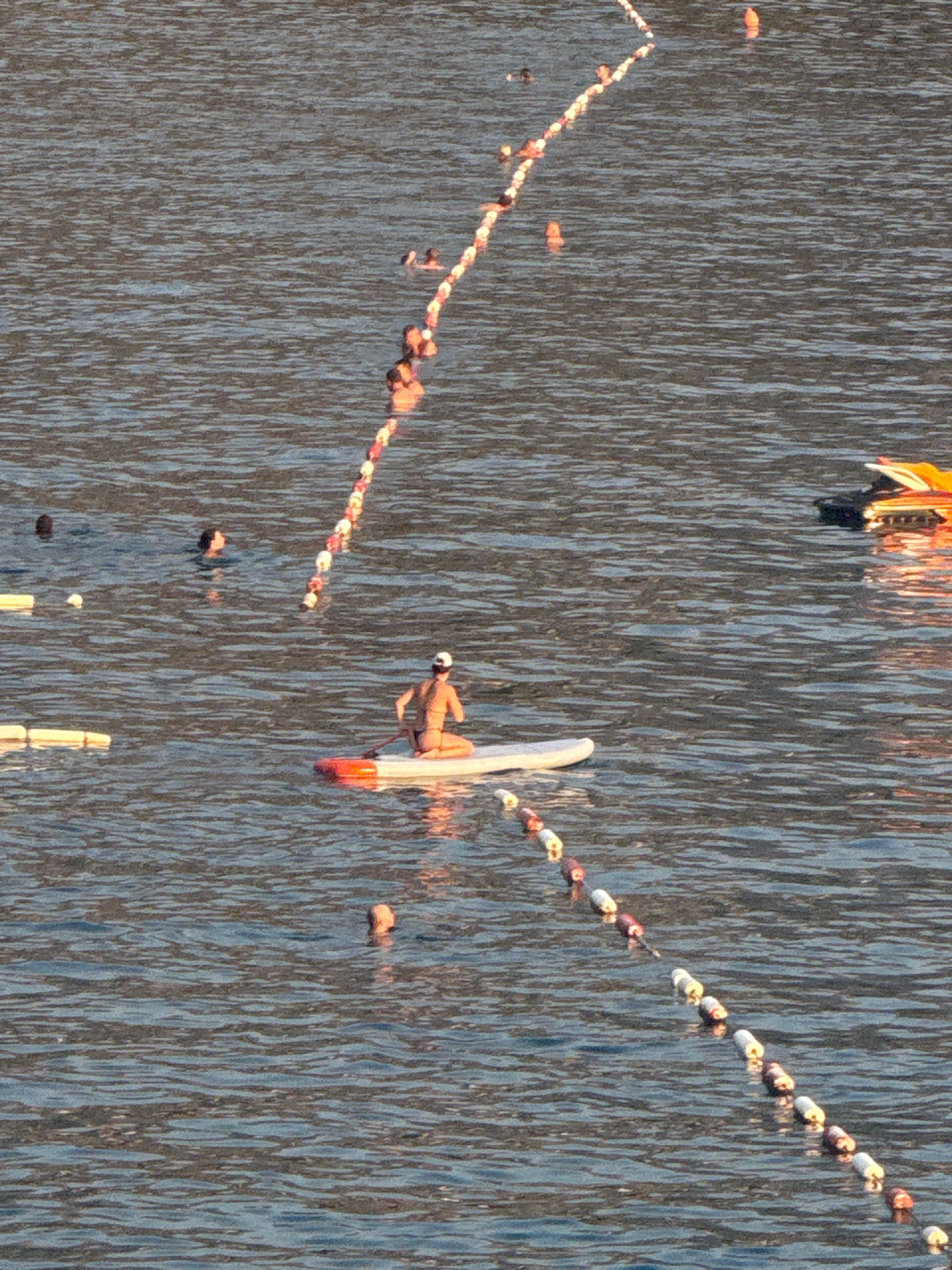 An aerial view shows a section of the water at Kamenovo beach in Montenegro. A person is visible on a stand-up paddleboard, paddling with a single-bladed paddle. The person is wearing a hat and swimwear. In the background, several other individuals are in the water, swimming or floating. A rope with buoys separates a portion of the water. In the upper right corner of the frame, a bright orange object, possibly a small boat or inflatable toy, is partially visible. The water is calm with a rippled surface reflecting the sunlight. The seabed is not visible due to the depth of the water.