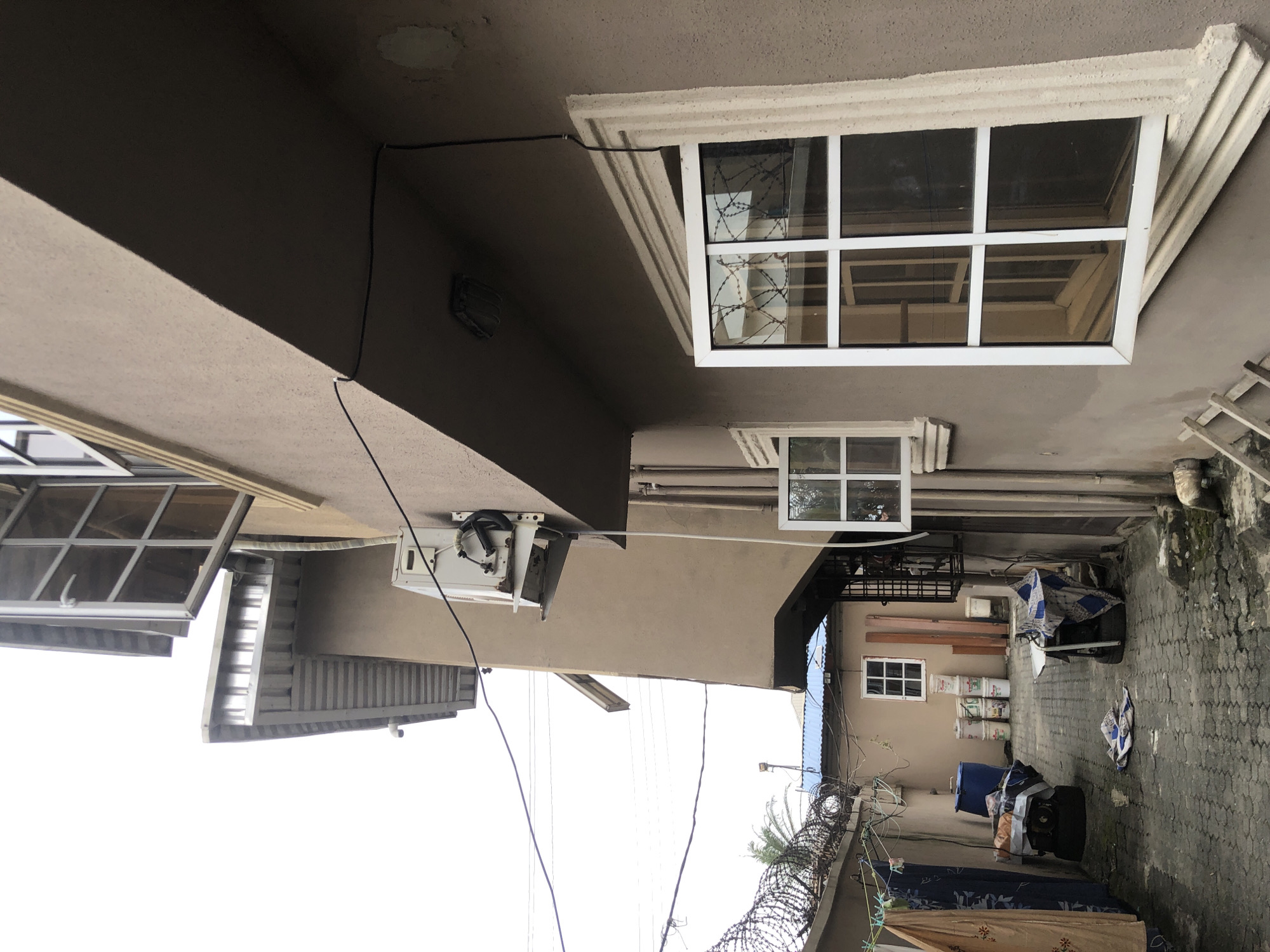 The image depicts a narrow outdoor alleyway between two buildings in Ado, Nigeria. The alleyway is paved with interlocking bricks and is somewhat cluttered with various items. On the left side, laundry is hanging on a clothesline, and there are several large blue plastic containers and other miscellaneous objects on the ground, some covered with fabric. In the background of the alley, there is a small building with a blue roof and a window. A barbed wire fence runs along the top of the wall separating the alley from another area.
The building on the right side of the alley is a multi-story structure with a stucco exterior painted a muted brownish-tan. It features several windows. One large window with white framing and multiple panes is prominently visible in the foreground, reflecting the barbed wire fence and the opposite building. Another smaller window is located above and to the left of the larger one. An air conditioning unit is mounted on the exterior wall of this building. Electrical wires and conduits are visible running along the exterior of the building. The overall scene suggests a residential or commercial area with a practical, somewhat utilitarian aesthetic. The sky is overcast.