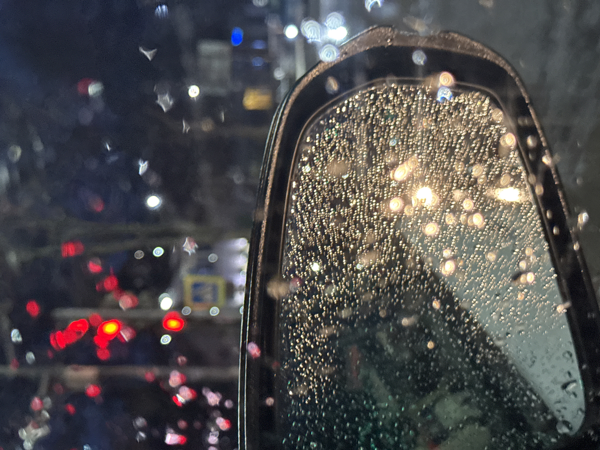 A photograph taken from inside a vehicle, likely a car, at night during a rainstorm. The primary focus is the passenger-side rearview mirror, which is covered in raindrops. The droplets reflect the diffused light from unseen sources, creating numerous bright spots.
Outside the mirror, through the rain-streaked window, blurred lights suggest a nighttime urban environment. Red and white lights are prominent, possibly from traffic signals or car headlights. Other blurred lights are visible and could be streetlights or lights from buildings. The city is not explicitly identified.