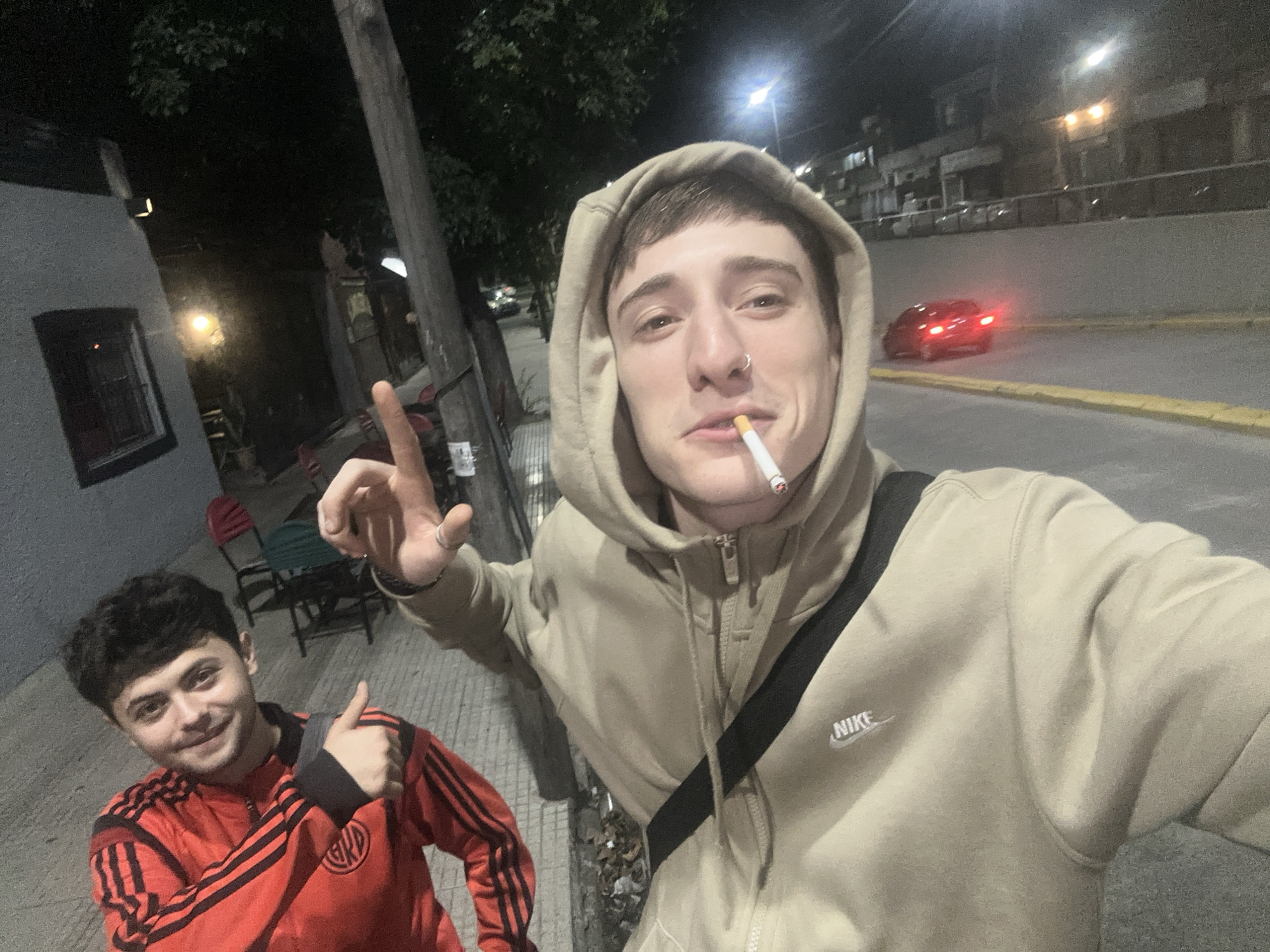 Two young men are outdoors at night in Villa Adelina, Argentina. The man closest to the camera wears a tan hooded Nike sweatshirt, a black shoulder bag, has a cigarette in his mouth, and holds up his index finger. The second man, slightly behind, wears a red and black striped jacket and gives a thumbs-up. A car with its rear lights on is visible on a street with a yellow curb. A building and trees are present in the background, illuminated by street lights. A grey building with a window is to the left with red chairs on the sidewalk.