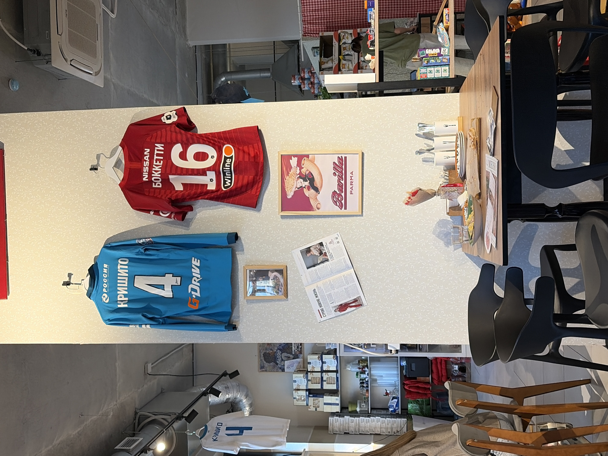 The image depicts a wall adorned with sports memorabilia and food-related items, alongside a dining area, likely within a cafe or restaurant in Izhevsk, Russia, given the Cyrillic text on the items. The wall features three framed sports jerseys, two red and one blue, all with Cyrillic lettering and numbers. A framed Barilla pasta advertisement and a newspaper with a picture of a person is also mounted on the wall. Below the wall, there is visible shelving filled with various food items, and lower down, another sports jersey hanging. The area transitions into a dining space with a wooden table and black chairs.