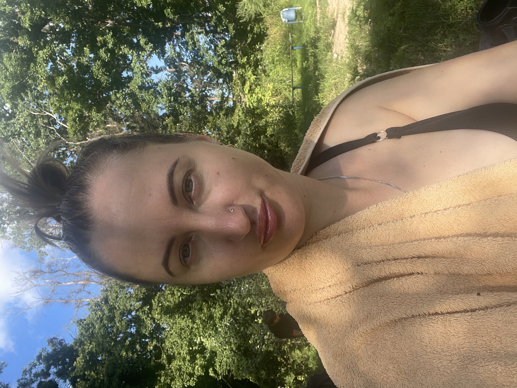 A woman is pictured in a close-up shot, her face and upper body taking up most of the frame. She has her hair pulled up in a bun and is wearing a dark-colored bikini top, with a tan towel draped over her shoulder. She is looking directly at the camera with a slight smile. In the background, lush green trees and a blue sky are visible, suggesting an outdoor setting, possibly a park or natural area. A faint shadow suggests the presence of another person behind her on the left, and further in the background, there appears to be a wooden bench and a blue trash can, indicating a public space. The lighting suggests it is daytime, likely sunny given the bright sky and shadows. The location is identified as Zarichne, Ukraine.