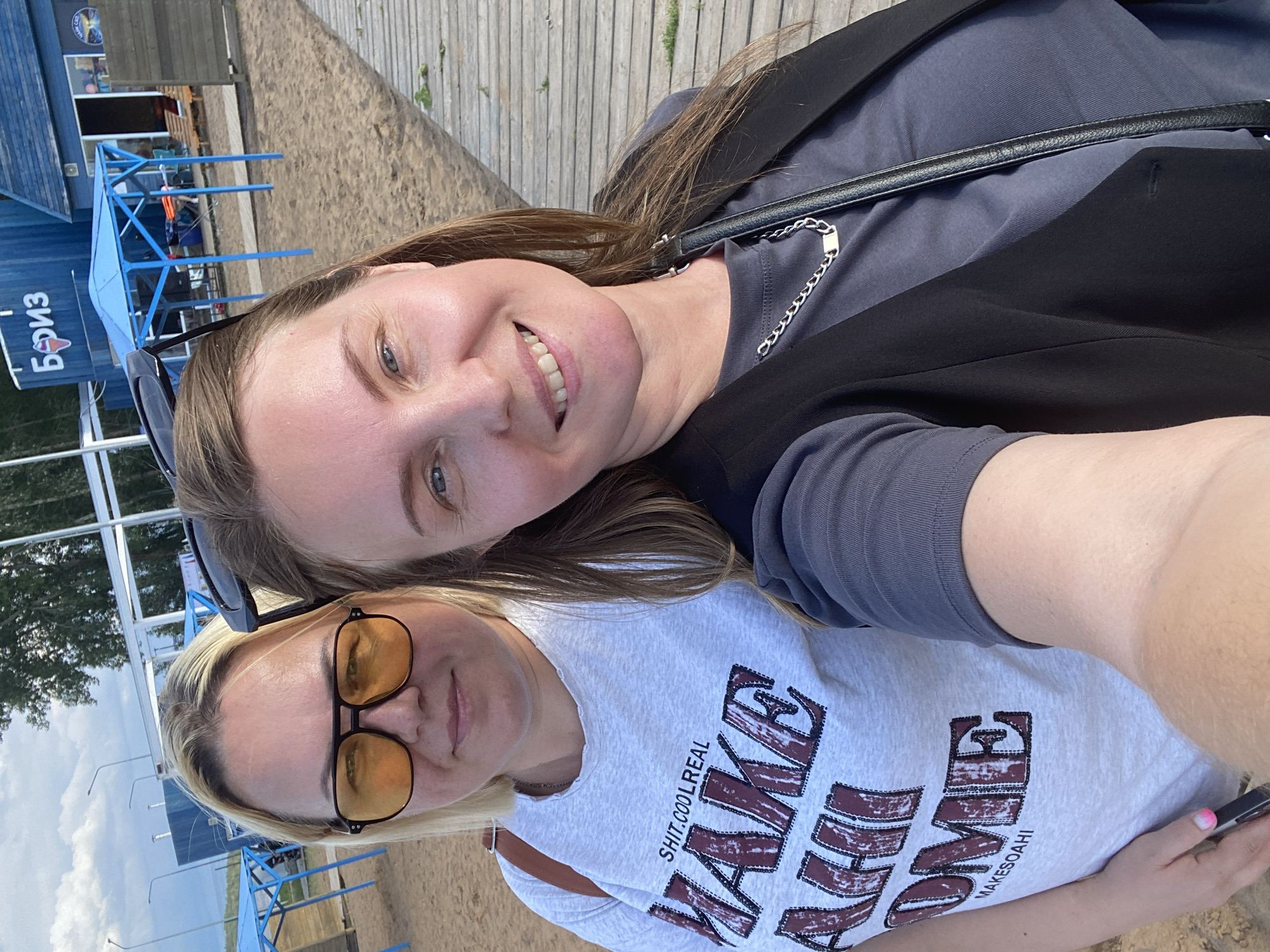 Two women are taking a selfie outdoors. The woman on the left has blonde hair and is wearing sunglasses and a t-shirt with text that reads "SHIT.COOLREAL MAKE AHJI OME MAKEOAHI". The woman on the right has brown hair and is wearing a dark vest over a grey t-shirt and a silver chain necklace. They are standing on a wooden boardwalk next to a sandy area. In the background, there is a blue building with Russian text that reads "БОРИЗ" (Boriz) and a blue canopy structure. This appears to be a cafe or beach facility, possibly located on the Kama River embankment in Chaikovsky, Russia. The sky is partly cloudy.