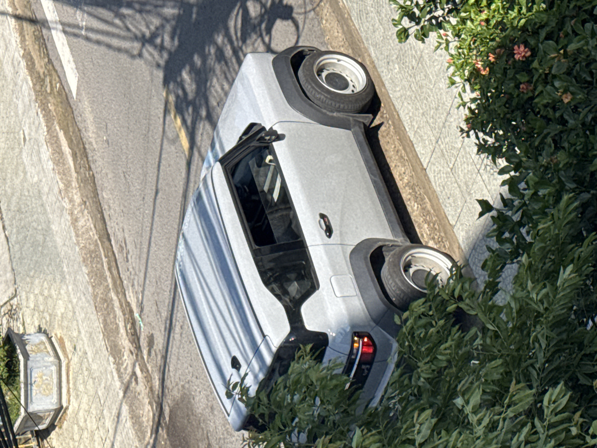 An aerial view depicts a silver car parked partially on a sidewalk adjacent to a street, partially obscured by foliage. The location is Da Lat, Vietnam.