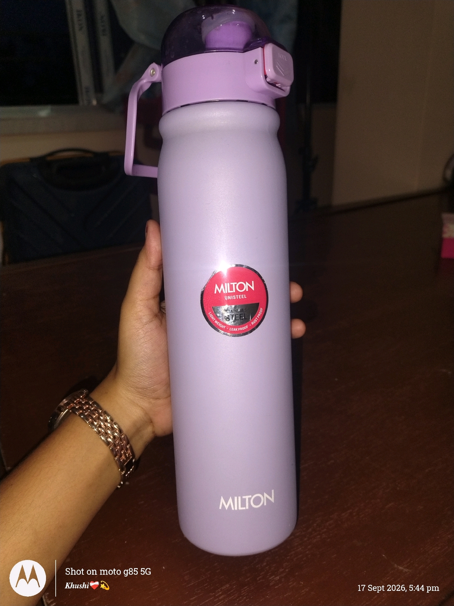 A pale lavender Milton Unisteel water bottle is held in a person's right hand. The bottle has a light purple lid with a flip-up spout and a purple carrying strap attached to the side. A red circular logo with the Milton brand name is visible on the bottle. The person is wearing a rose gold metal band watch on their left wrist. The background shows a dark brown wooden table surface. The bottom left corner of the image displays the Motorola logo and text indicating "Shot on moto g85 5G Khushi," and the bottom right corner shows the date and time "17 Sept 2026, 5:44 pm." There is no identifiable location in Barrackpore, India, or any city.