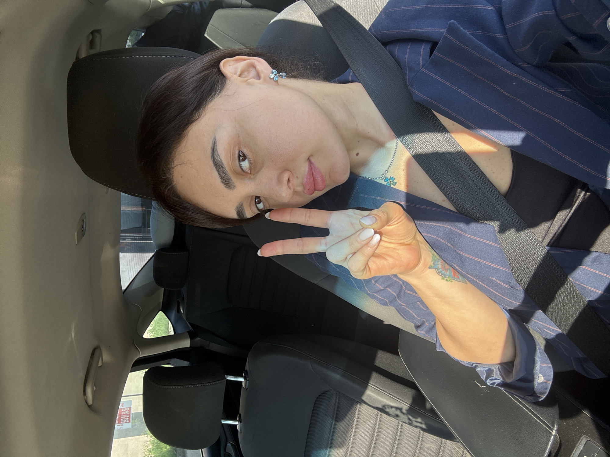 A woman is seated in the driver's seat of a car, wearing a navy pinstripe blazer over a black top, secured by a seatbelt. She is making a peace sign with her right hand. Her dark hair is pulled back. She has a light blue flower earring and a silver necklace with a blue pendant. A butterfly tattoo is visible on her inner right forearm. Outside the window, a building with posters on the side of it can be seen. The exact location in the city of Tbilisi, Georgia cannot be determined from the given information.