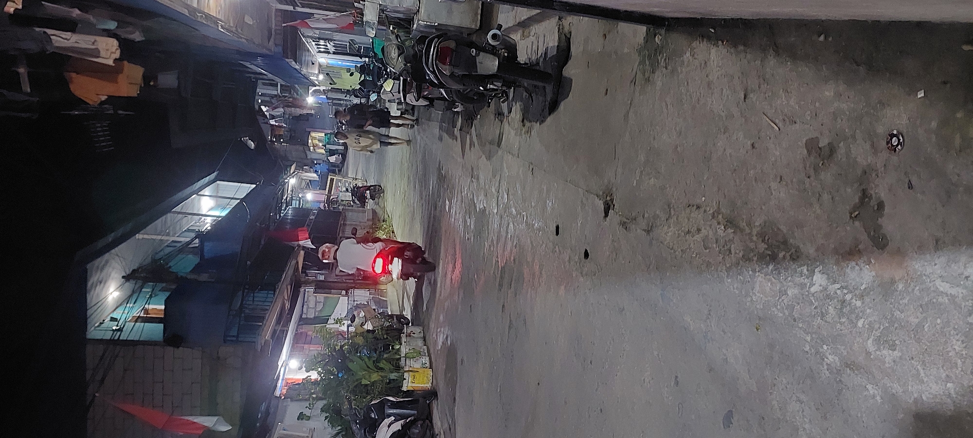 An outdoor nighttime vertical shot depicts a narrow, paved alleyway in Jayapura, Indonesia. The alley is flanked by multi-story buildings and appears to be a residential or commercial area. The lighting suggests it is late at night or very early morning, with artificial lights illuminating parts of the scene.
In the foreground, a person is riding a red motorcycle away from the camera. The motorcycle's taillight is illuminated, casting a red glow on the pavement. The rider is wearing a white shirt and a helmet. To the left of the motorcycle, several other motorcycles are parked. A small potted plant with lush green leaves is visible next to the parked motorcycles.
Further down the alley, two individuals are walking towards the motorcycle. One person is wearing a light-colored hooded sweatshirt and dark pants, while the other is wearing a dark t-shirt and dark shorts. More parked motorcycles are visible on the right side of the alley.
The buildings lining the alley exhibit varying architectural styles and states of repair. Some have visible brickwork and balconies, while others appear to have corrugated metal roofs. Laundry is hanging from lines strung between buildings. Indonesian flags, red and white, are visible on some of the buildings, suggesting a festive or nationalistic occasion.
The pavement is uneven and appears to be concrete. Scattered debris, including small pieces of wood and what looks like a discarded lid or cap, are present on the ground. The overall impression is a densely populated urban environment, likely a slum or informal settlement, with vibrant street life even at night.