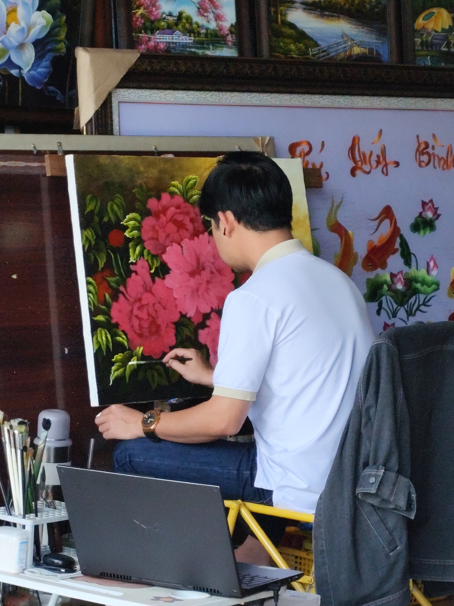 The image shows a male artist painting a large canvas with vibrant pink peonies. He is wearing a white polo shirt with a beige collar and dark blue jeans. He is holding a paintbrush in his right hand and appears to be in the process of adding detail to the flowers. To his left, a laptop is open on a white table, and to his right, a dark gray jacket is draped over a yellow easel. In the background, a wall is decorated with embroidered art featuring orange goldfish and lotus flowers. The overall scene suggests an art studio or a market stall, possibly in Lam Vien - Da Lat, Vietnam, given the common artistic displays in such locations. Other art pieces are visible in the background, including paintings of lotuses and a landscape scene. Brushes and art supplies are neatly arranged on a white shelf to the left of the artist.