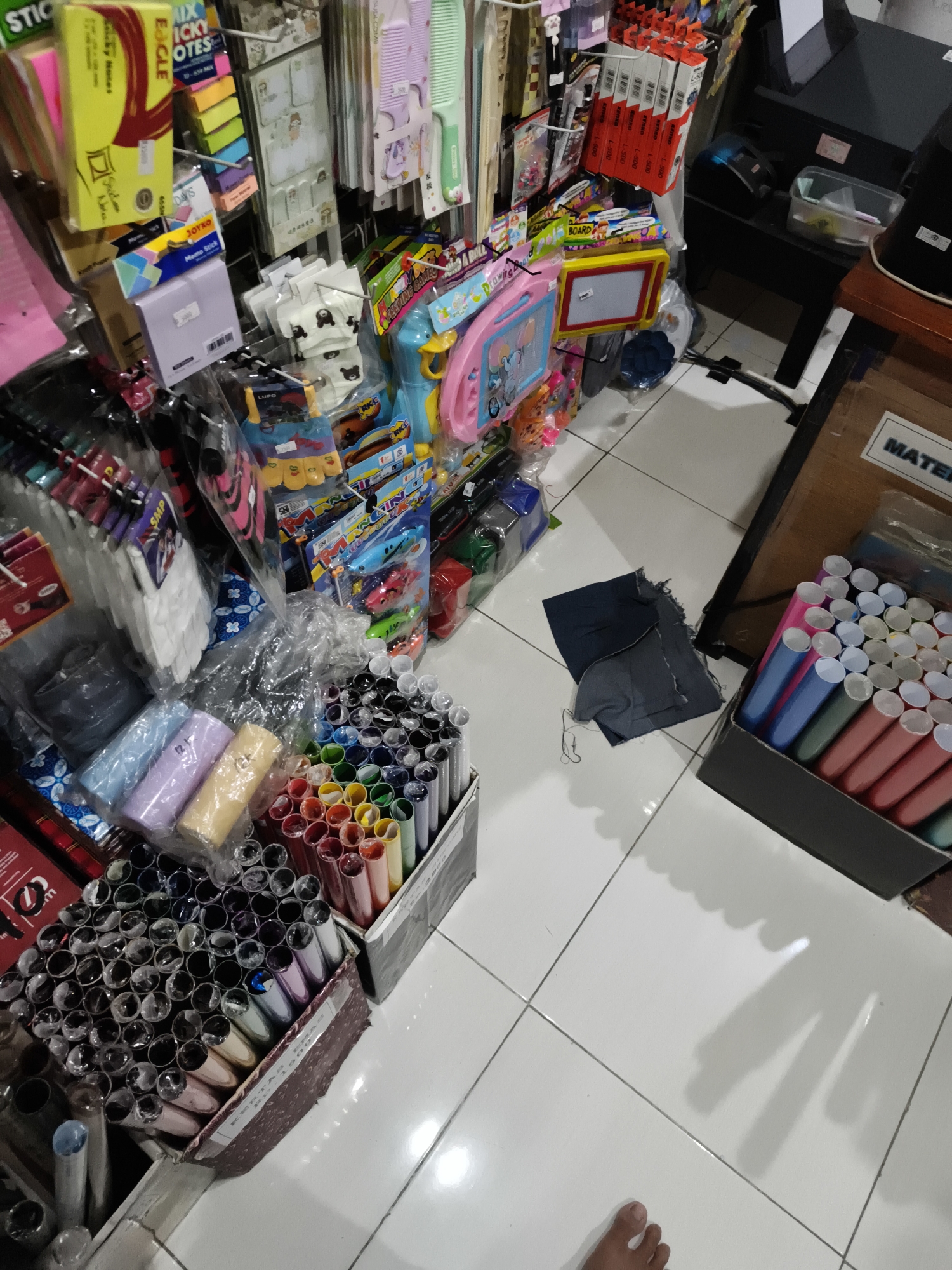 This is a high-angle, medium shot of a retail display area, likely a store that sells stationery and art supplies. The scene is brightly lit, suggesting an indoor environment, possibly during the daytime. The floor is tiled in white.
The primary subjects are an abundance of colorful objects, including rows of pens or markers in various shades, wrapped individually in plastic. These are neatly arranged in cardboard boxes. There are also various packaged stationery items, such as sticky notes, combs, and what appear to be small drawing boards. Some items are displayed hanging from a rack system. Notably, there is a pile of dark fabric, possibly denim, on the floor. In the lower right corner, a person's foot is visible, indicating a person is present in the scene, though only their foot is in frame.
There is visible text in various languages, including "STICKY NOTES," "EAGLE," "JOYKO," "Memo Stick," "RKC," "MATER," and numbers like "500." The location context provided indicates this is Sidorejo, Indonesia. The overall impression is one of a well-stocked shop with a wide variety of colorful goods for sale. No specific activity is clearly taking place, but the arrangement suggests a commercial setting where items are displayed for purchase.