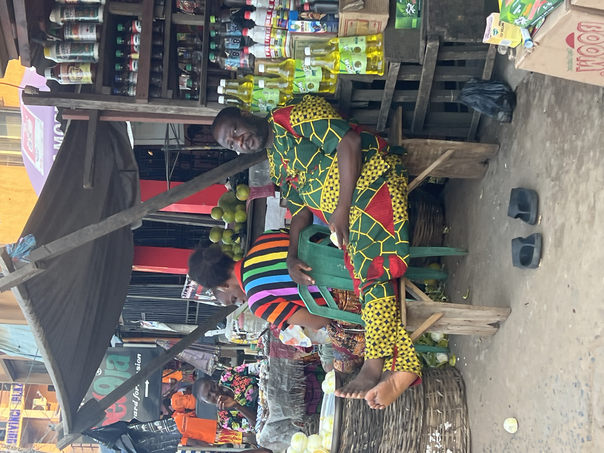 In Nkpor, Nigeria, a man in a brightly colored green and yellow patterned outfit sits on a wooden stool in front of a market stall. His legs are crossed, with one foot resting on the stool. To his right, shelves are stacked with various bottles, some clear with yellow liquid, others with red and white labels. In the background, a woman in a floral shirt is seated at a table with goods laid out, and another person wearing a striped shirt sits on a green plastic chair. The market stall has a dark tarp overhead, supported by wooden beams. The ground is paved with concrete, and several baskets are visible. In the foreground, two black flip-flops lie on the ground.