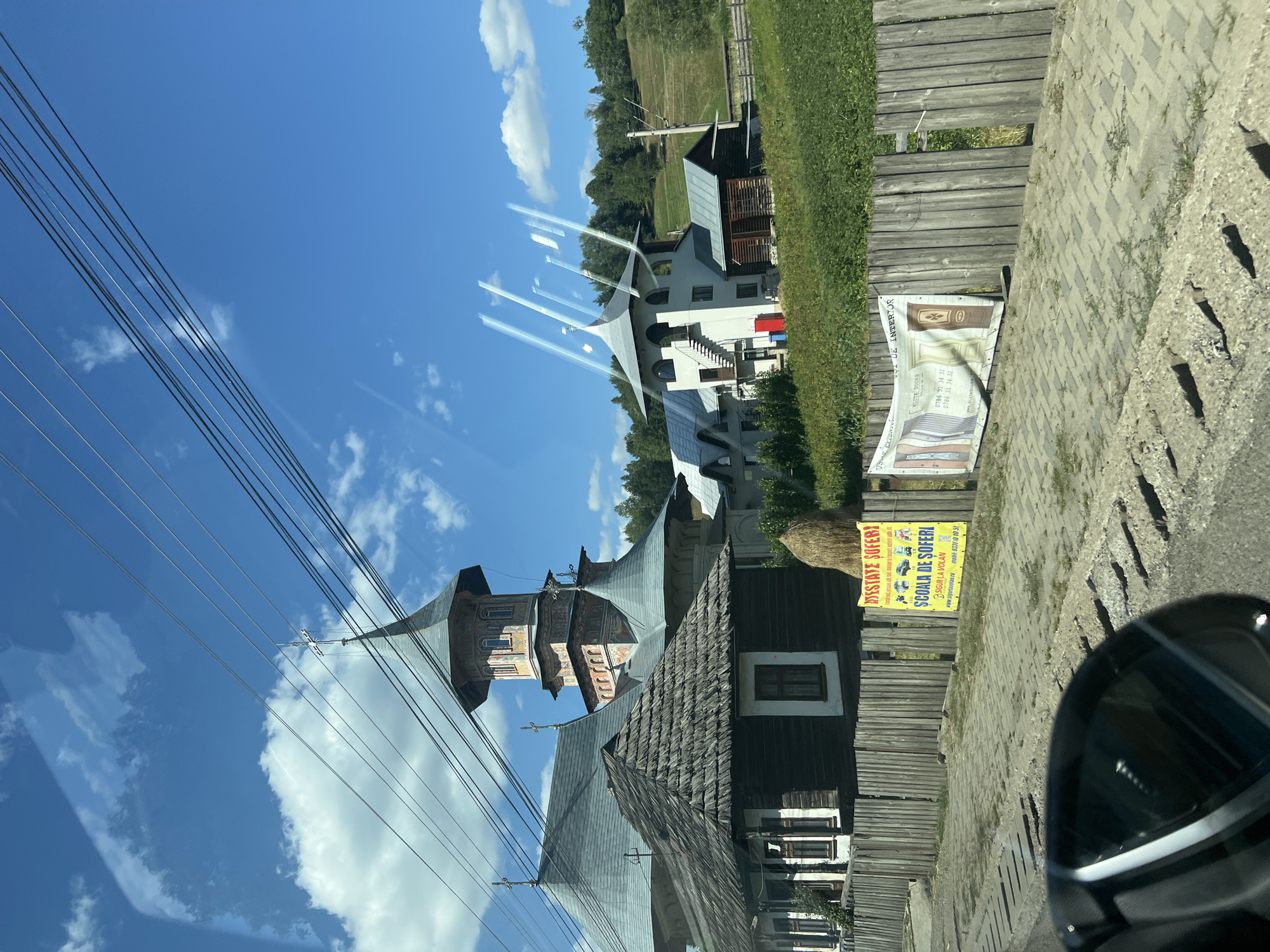 The image depicts a street scene in Molid, Romania, on a sunny day. The foreground shows a cobblestone street and a wooden fence. Beyond the fence, a traditional Romanian wooden house with a thatched roof is visible. Behind the house, a larger religious structure with multiple towers and a distinct architectural style, possibly a monastery or church, is present. The sky is blue with scattered white clouds. Several overhead power lines are prominent in the upper part of the frame. Two advertising banners are affixed to the wooden fence; one is yellow with text and images of cars, advertising a driving school, and the other is a white banner displaying various interior doors. The overall impression is of a rural or semi-rural setting in Romania.