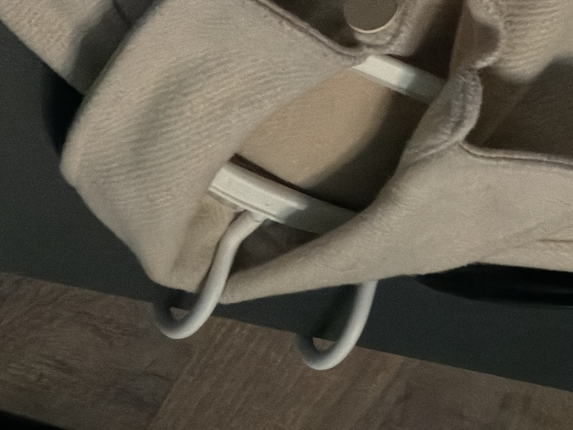 The image shows a close-up view of a light beige garment, possibly a coat or sweater, draped over a white hanger. The hanger itself is partially visible, with its curved arms supporting the fabric. The garment has a textured, ribbed pattern. In the background, a dark, possibly wooden surface with visible grain is seen on the left, and a dark gray surface is on the right. The overall impression is that of clothing stored or displayed indoors, potentially in a domestic setting or retail space. There is no visible indication of a specific location in the city of Sofia, Bulgaria.