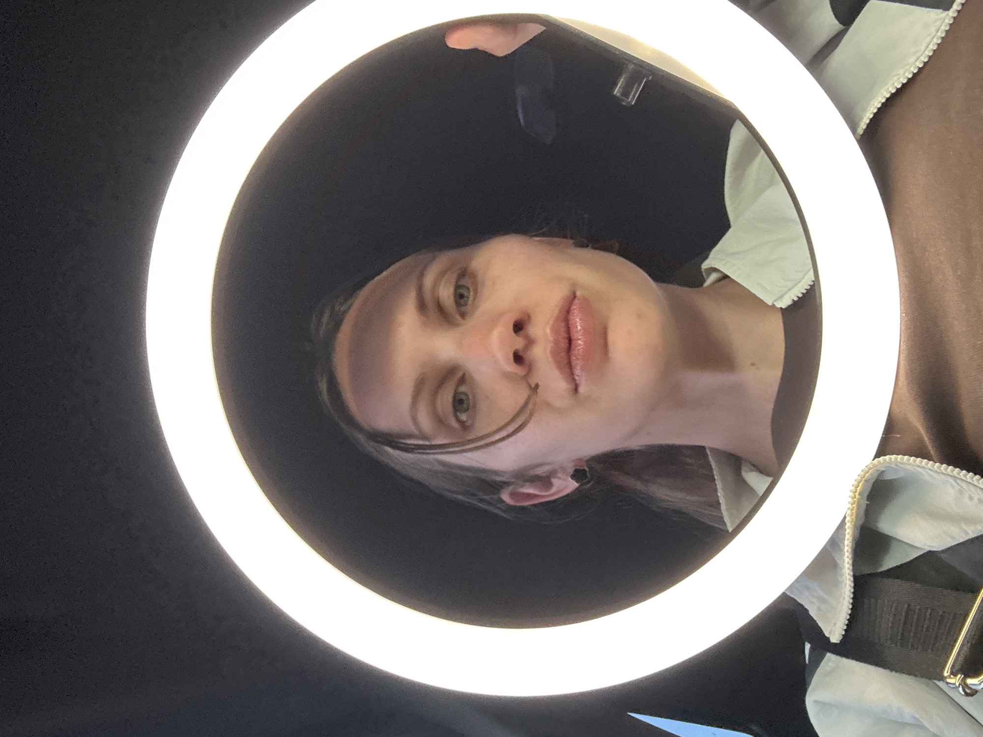 The image displays a close-up, rotated self-portrait of a person, likely female, dramatically illuminated by a bright, circular ring light. The subject's face is positioned centrally within the light's aperture, appearing to look downwards towards the camera lens. She has fair skin, light-colored eyes (appearing green or hazel), and a neutral to slightly pensive expression, with her lips slightly parted and exhibiting a glossy finish. A few strands of dark hair fall across her forehead and cheek, and a dark stud earring is visible in her left ear.
The prominent ring light casts a strong, even, and artificial glow on the subject's face. The area within the ring and immediately surrounding the face is dark, creating a stark contrast with the brilliant light. Faintly visible at the top edge of the ring light is a hand holding a smartphone, indicating the method of capture.
Partial views of clothing are seen at the periphery of the image. On the right side, a light-colored garment, possibly a jacket or shirt, is visible with a zipper and a decorative white, possibly scalloped, trim. A dark strap, potentially from a bag or another garment, is also present on the far right. The overall image is rotated, presenting the scene at an unusual angle. The artificial lighting and dark background suggest an indoor setting, possibly for a photoshoot or video recording. While the location context is Fethiye, Turkey, no specific visual cues within the frame confirm this. No discernible text is visible in the image.