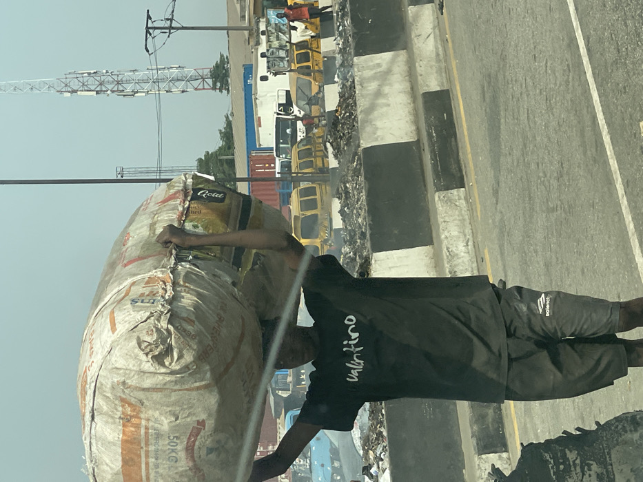 The image depicts an urban scene in Aqani, Nigeria, under bright daylight conditions. In the foreground, a dark-skinned male, appearing to be a young adult, walks from left to right. He wears a black short-sleeved t-shirt with "valentino" in white text across the chest and dark shorts with a "balenci" logo on the left thigh. He is carrying a large, irregularly shaped, off-white woven plastic sack on his head, supporting it with both arms raised and hands gripping the top edges. The sack bears faded red/orange text, including "50KG" and "ROLA."
The individual is on an asphalt road with visible yellow and white markings. To his right, a concrete barrier, painted with alternating black and white stripes, separates the road from an adjacent area. Behind this barrier, a significant pile of refuse, mixed with dark, burnt debris, is visible, with faint wisps of smoke indicating recent or ongoing combustion.
In the mid-ground and background, numerous vehicles are present. Several yellow mini-buses, typical of local public transport, are parked or moving behind the trash pile. Further back, various trucks, including those carrying red and blue shipping containers, are visible. A light-blue vehicle is partially seen on the far left. At least two other individuals are discernible in the far right background, one wearing an orange shirt.
The skyline includes a prominent tall, metal lattice telecommunication tower equipped with antennas, along with several utility poles and overhead lines. The overall environment suggests a densely populated area with active transport and waste management challenges.