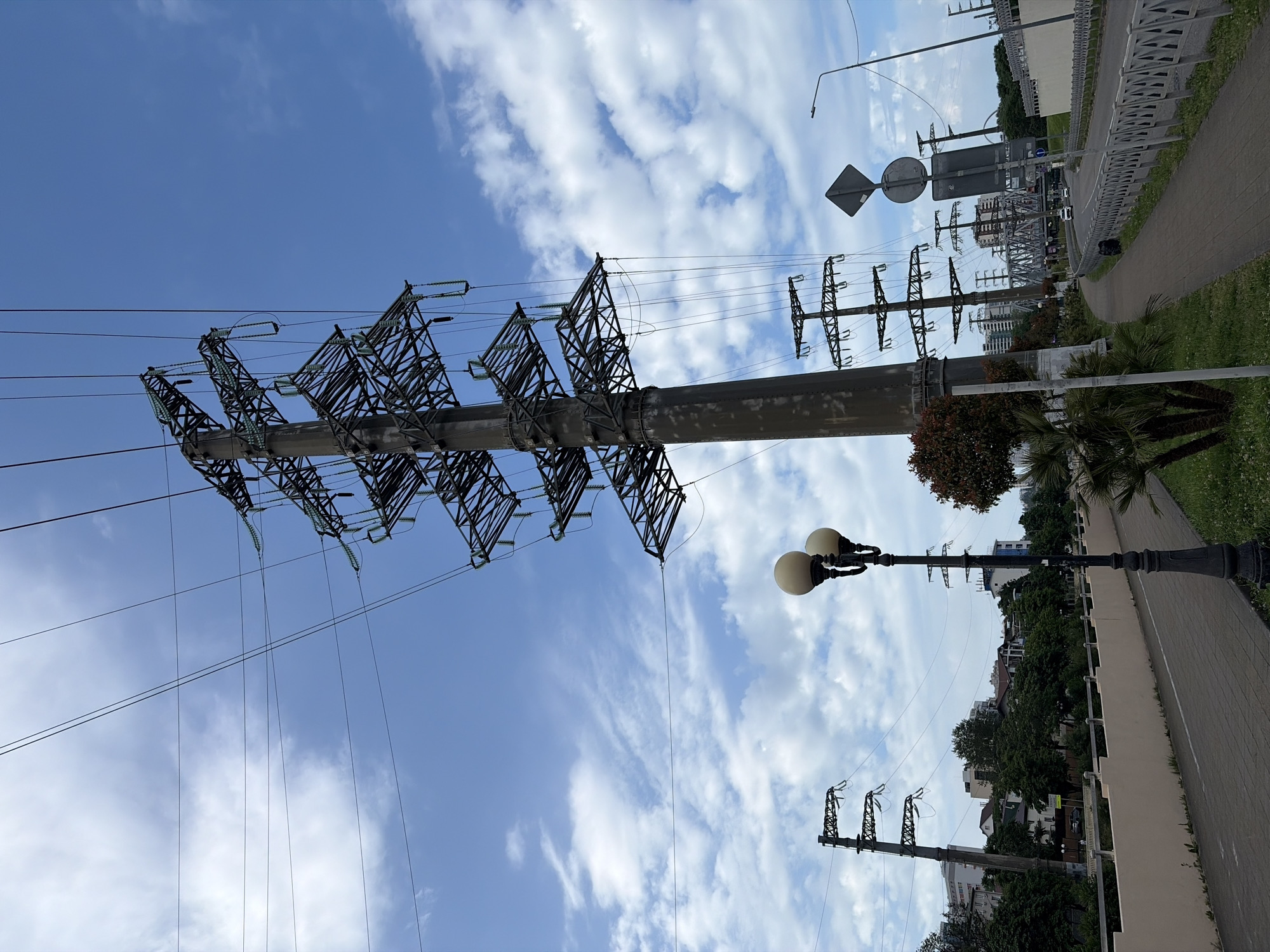 The image depicts a street scene in Sochi, Russia, characterized by a prominent power line pylon dominating the foreground. The pylon, made of metal lattice, extends upwards, supporting multiple electrical wires against a backdrop of a blue sky with scattered white clouds.
To the left of the pylon, a two-globe lamppost stands on a paved walkway. Further to the left, another, smaller power line pylon is visible, along with buildings and lush green trees lining the background. The walkway appears to be part of a pedestrian path or promenade, bordered by a light-colored wall and a strip of grass with some palm trees.
The right side of the image shows a road with a few cars and a metal fence. In the distance, a bridge can be seen, and more buildings are scattered throughout the urban landscape. The overall impression is that of an urban environment where infrastructure, including power lines and roadways, integrates with natural elements like trees and sky.