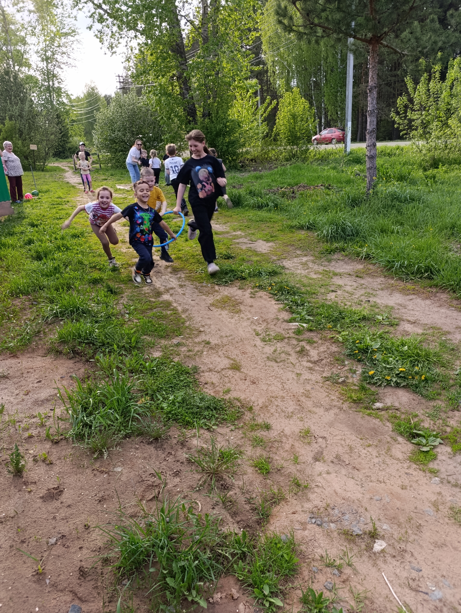 The image depicts a group of children playing outdoors on a dirt path in what appears to be a residential area in Chaikovsky, Russia. The children are running and appear to be participating in a game involving a blue hoop. An older girl leads the group.
There is an older woman standing near a building on the left side of the image. A few adults and other children are visible in the background near some trees.
The path is surrounded by grass and foliage, suggesting a park-like or semi-rural environment. A road is visible in the background, with a red car on it. The presence of power lines in the distance also indicate an urban location.