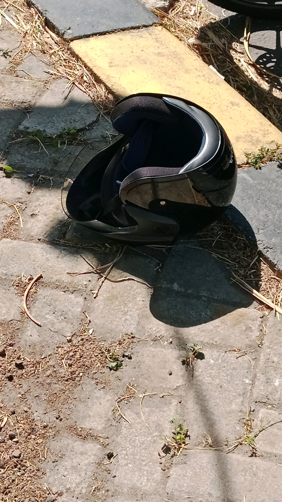 A black motorcycle helmet is lying on a concrete paved surface. The helmet is partially open, with the visor up, revealing the dark interior lining. The pavement is irregular, with some gaps filled with dry leaves and soil. A yellow concrete slab is visible in the upper right portion of the image, with a dark gray concrete slab next to it. The scene is outdoors and appears to be a sunny day, casting a shadow from the helmet onto the pavement. There is no indication of any people or any specific activity taking place. The location is described as being in the city of Sugihwaras, Indonesia.