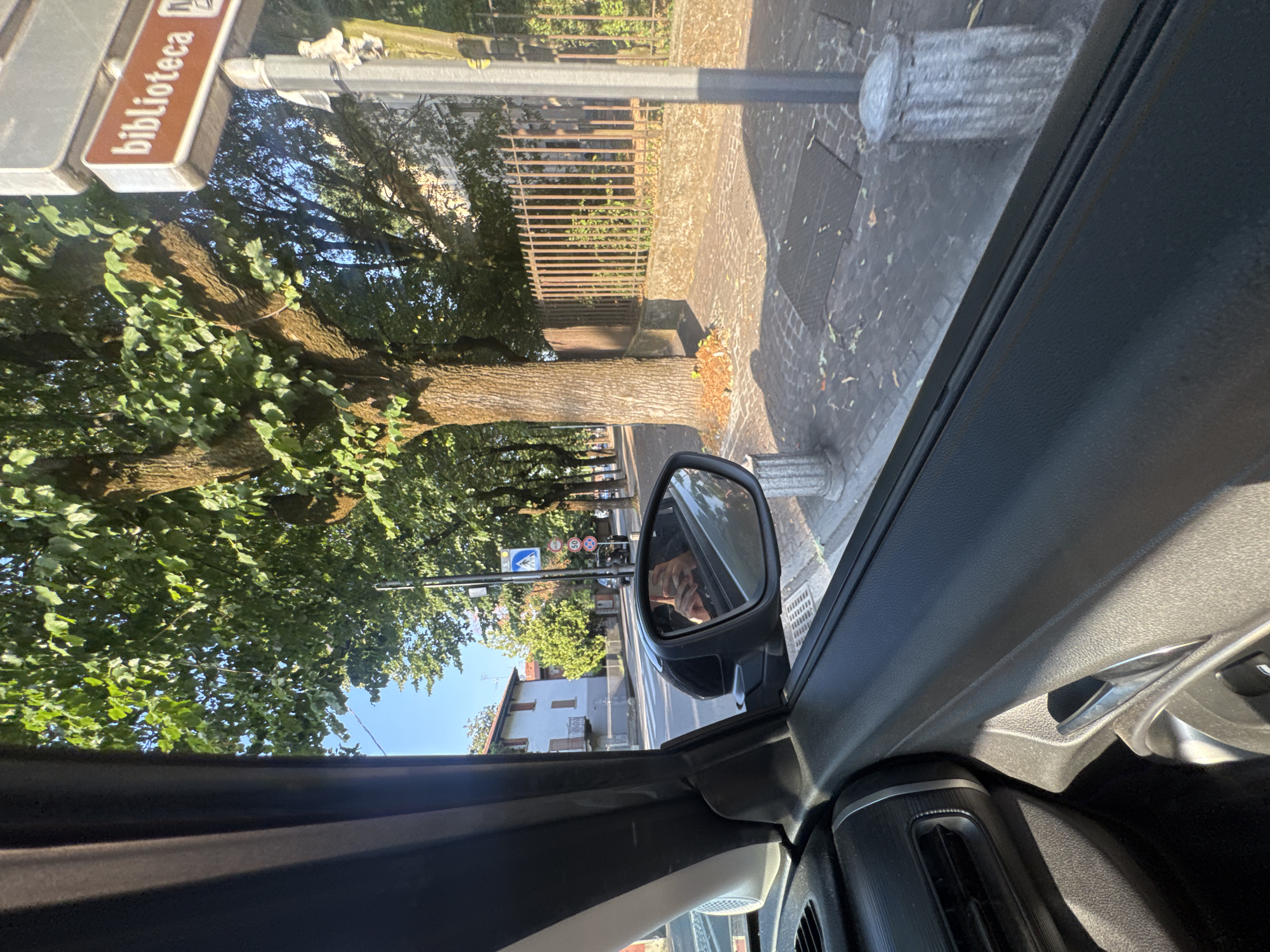 The image captures a view from inside a car, looking out the passenger side window towards a street scene in Chiari, Italy. A large tree with green foliage dominates the midground, casting shadows on the cobblestone sidewalk. To the right, a street sign reads "biblioteca." In the distance, a building with a light-colored facade and brown shutters is visible. A metallic fence runs along the sidewalk. The side mirror reflects a person holding a smartphone and taking a photo. The car's interior, including the door panel, air vent, and door handle, is visible in the foreground. The sky is clear and blue.