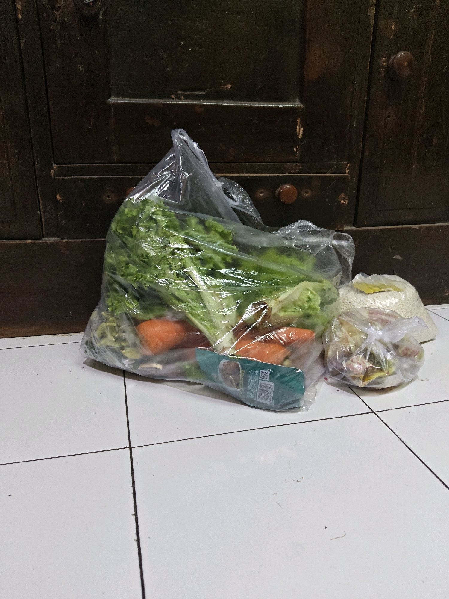 The image displays grocery bags filled with fresh produce and rice placed on a tiled floor in front of a dark wooden cabinet. The largest bag contains lettuce and carrots. To the right, there are two smaller bags; one appears to contain rice, and the other contains various smaller food items. The background is a dark, aged wooden cabinet with visible hardware and wear. The lighting is dim, suggesting an indoor setting, possibly a kitchen or storage area in Sumberwono, Indonesia, given the context. There are no people present in the image, and no actions are being taken. The scene is static.