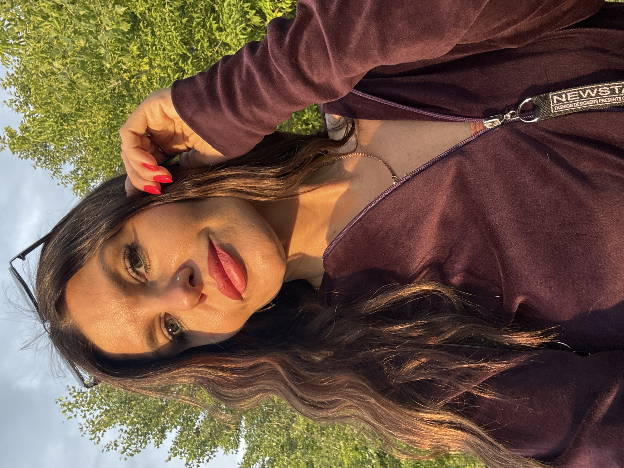 This is a close-up, vertically oriented photograph of a woman's face and upper torso, taken outdoors in Chaikovsky, Russia. The woman has long, wavy brown hair and is wearing a dark purple zippered top, possibly a hoodie or sweatshirt, which has a tag on the zipper pull that reads "NEWSTAY FASHION DESIGNERS PRESENTS". She has bright red nail polish on her fingers, and her left hand is resting on her head. She is wearing makeup, including lipstick and mascara, and has on dark-rimmed glasses. The lighting suggests it is late afternoon or early evening, with warm sunlight illuminating her face and casting shadows. Behind her, a lush green tree is visible against a bright sky, indicating a pleasant, possibly warm, day. The overall mood appears relaxed and natural.