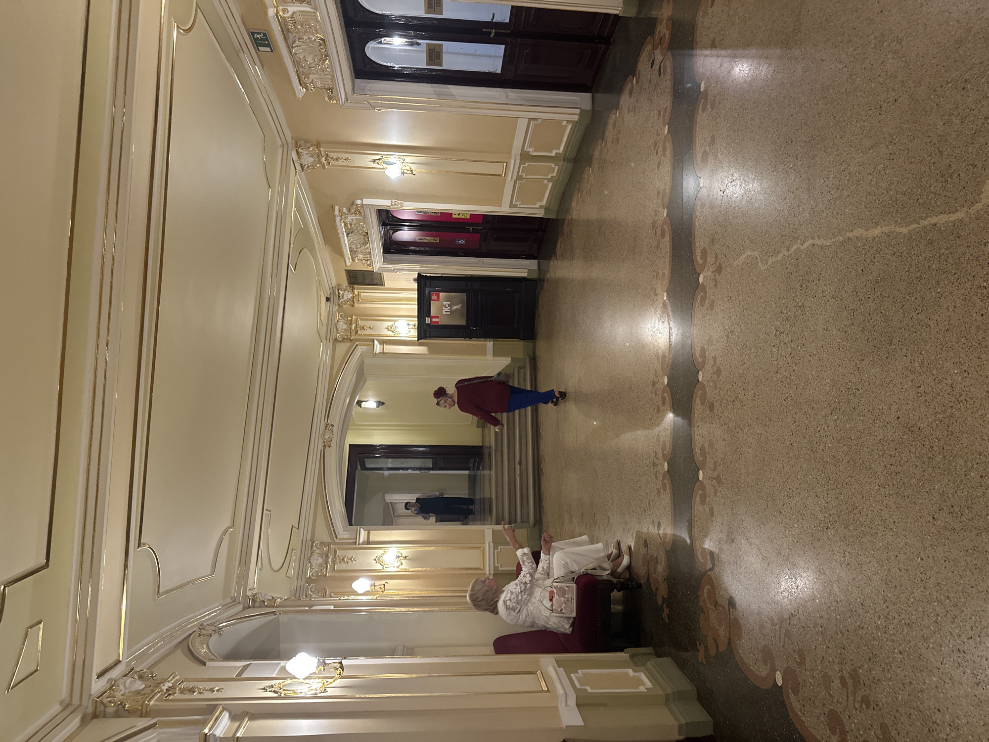 The image depicts a richly decorated interior hallway, likely within a public building such as a theater or opera house, given the ornate gold trim, moldings, and patterned terrazzo flooring. The scene is illuminated by warm interior lighting, suggesting it is evening or indoors.
In the foreground, two women are seated on a plush red armchair. One woman, wearing a white, intricately patterned top and white pants, is looking upwards and gesturing with her left hand. She has a light-colored handbag on her lap. The other woman, partially obscured, appears to be wearing a similar white outfit and is facing away from the camera.
Further down the hallway, a person dressed in a maroon jacket and blue pants walks away from the camera, heading towards a set of doors. In the background, several dark wooden doors with glass panels are visible, some with "EXIT" signs. There is also a glimpse of stairs leading upwards, with another person visible on the landing.
The overall impression is one of elegance and historical grandeur. The lighting creates a warm and somewhat subdued atmosphere, and the presence of people suggests it might be during a performance or an event. No specific time of day or weather cues are evident, beyond the interior setting. The location context is Odesa, Ukraine.