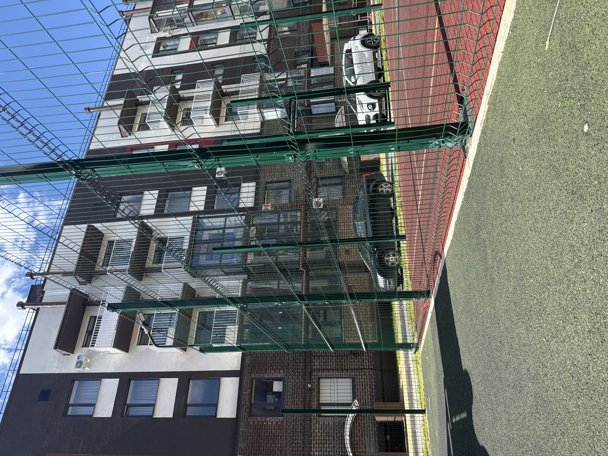 This is a vertical, eye-level shot of an apartment building and a sports field, taken on a sunny day in Viazniky, Russia. The building is tall and modern, with a dark brown and white facade, featuring numerous windows and balconies. A green metal fence runs across the foreground, partially obscuring the view of the building and a sports field with a red running track and a green artificial turf area. Two cars are visible parked in front of the building, one white and one dark. The sky is blue with scattered white clouds. The overall impression is one of an urban residential area with recreational facilities. There is no visible text in the image.