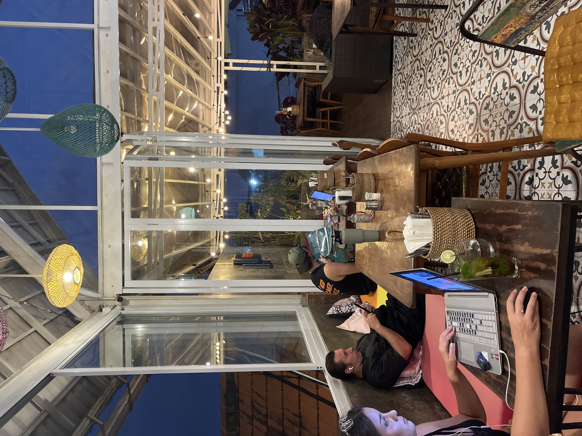 The image depicts an indoor seating area of a cafe, likely in North Kuta, Indonesia, at dusk. The scene is brightly lit by overhead string lights and decorative lamps, with the dim twilight visible through the glass walls. Three individuals are present. In the foreground, a woman in a black top looks towards a laptop on a wooden table, her hands positioned on the keyboard. Beside the laptop are a glass with a drink and lime, a woven basket with napkins, and what appears to be coffee beans. Another person, a man wearing a black tank top and a green cap, is seated at the same table, looking at a book or papers. Further back, a man in a black shirt is lying down on a pink cushion, engaged with his phone, wearing white earbuds. The cafe has a modern yet cozy ambiance, with patterned tiles on the floor and wooden furniture. The overall mood appears relaxed and casual, suggesting a typical evening at a cafe.