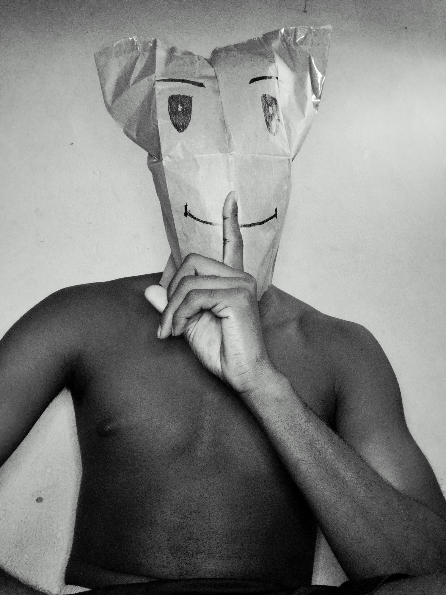 A medium shot, black and white, outdoor photograph depicts a person wearing a paper bag over their head. The bag has cartoonish eyes and a mouth drawn on it, and a finger is pressed to its lips, indicating silence. The person's bare torso is visible, with arms crossed in front of them. The background is a plain, light-colored wall. The person's skin tone is dark. The image appears to be taken in a studio or indoor setting, with a plain wall as the backdrop. There are no visible elements that place the image in a specific city or outdoor location.