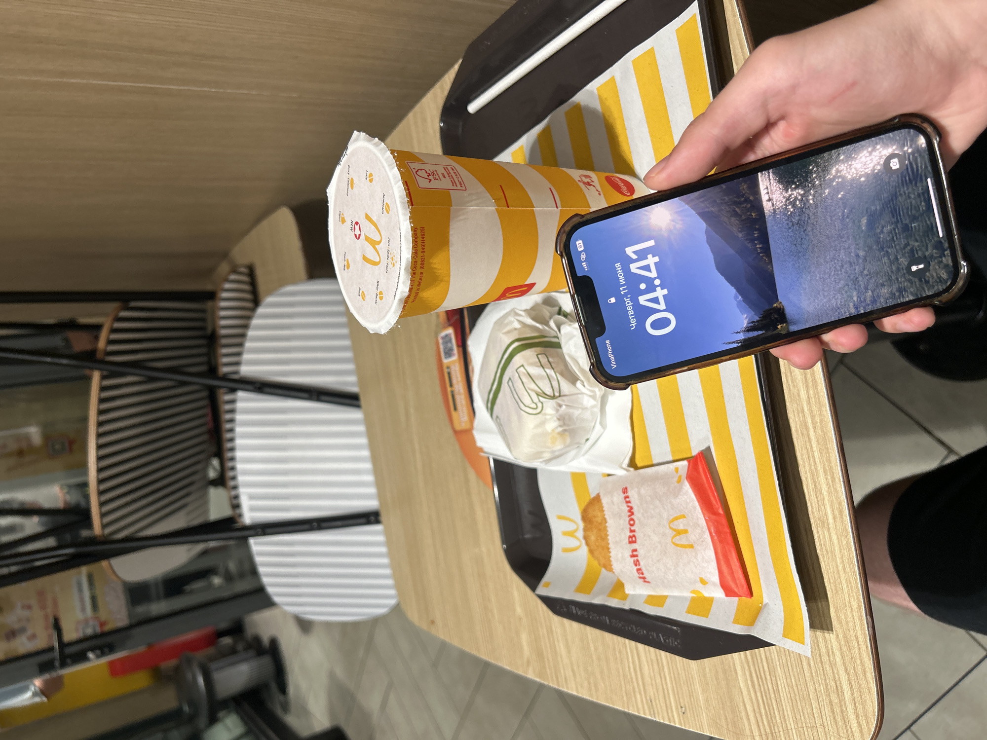 The image depicts a tray of McDonald's food on a wooden table. On the tray, there is a paper bag containing "Hash Browns" and a wrapped burger. To the right of the tray is a large yellow and white striped McDonald's cup with a lid. In the foreground, a person's hand holds a smartphone displaying the time "04:41" and the date "Thursday, June 11". The background shows blurry elements of a McDonald's restaurant interior with seating and decor. The overall scene suggests a meal being consumed or about to be consumed in a fast-food establishment. It is stated that this location is in Thủ Đức, Vietnam.