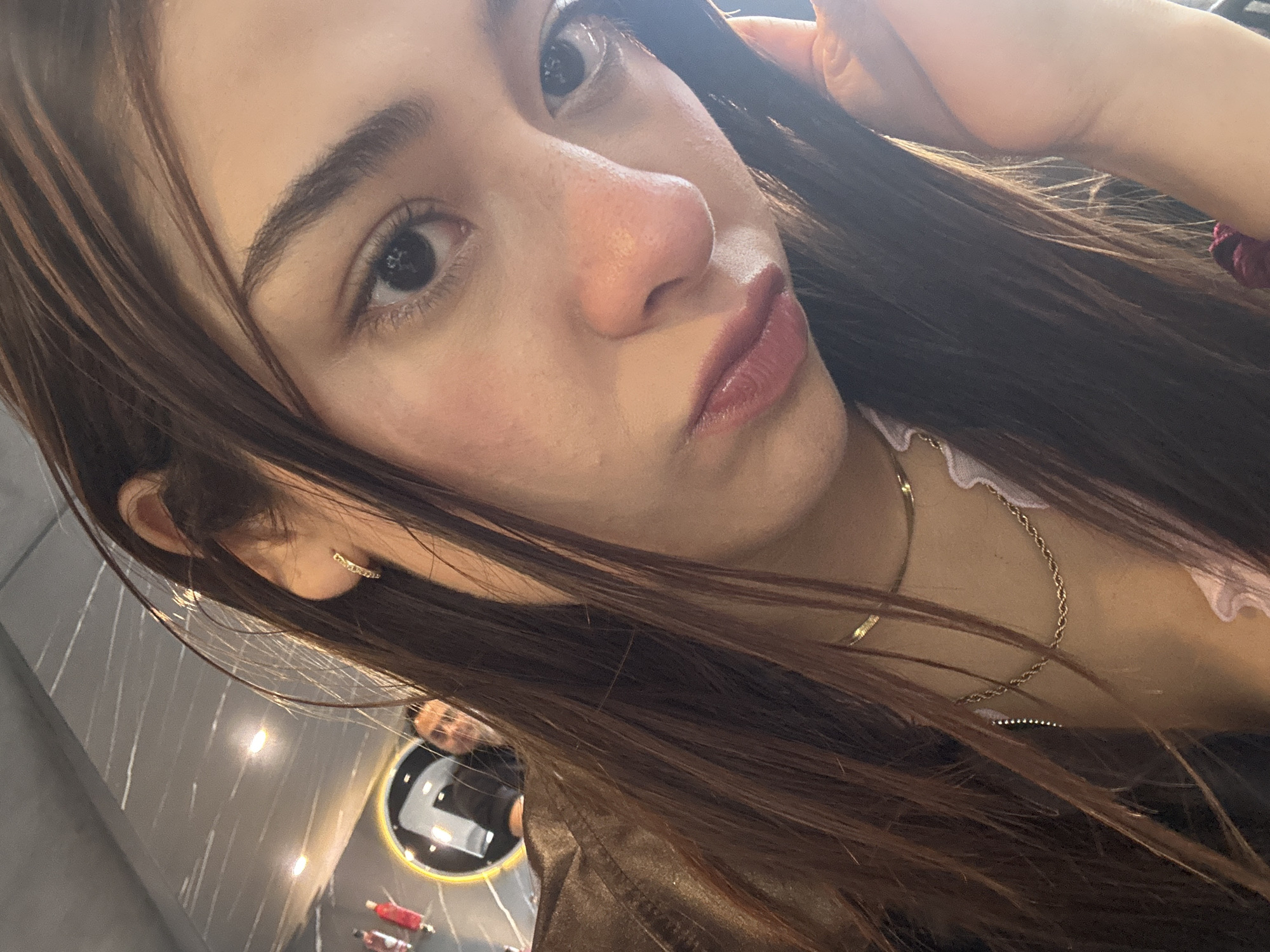 An indoor photograph, likely a selfie, features a young adult female in San Andrés Cholula, Mexico. The subject, positioned centrally, has long, straight, dark brown hair that falls around her face and shoulders. Her head is tilted slightly, and her dark eyes are directed towards the camera. Her complexion appears fair to medium, with visible pores and some redness on her nose and cheeks. Her lips are full, enhanced with a reddish-pink cosmetic, and she exhibits a slight pout.
She wears a small, gold-toned hoop earring in her visible left ear. Around her neck are two gold-toned necklaces: an upper, flat snake chain and a lower, twisted rope chain. Her attire includes a brown, likely leather, outer garment and a light-colored, possibly white or pink, ruffled garment underneath. A portion of her right hand is visible in the lower right, with a dark red scrunchie on her wrist or holding her hair.
The background reveals an indoor commercial setting, possibly a restaurant or cafe. A dark gray ceiling with linear white patterns and warm-toned recessed light fixtures is visible. In the upper left background, a circular mirror with a bright yellow rim reflects the upper torso and head of a male individual with short dark hair, looking towards the camera. Adjacent to the mirror, two small, red, bottle-like objects are partially visible on a surface. The scene is illuminated by ambient artificial light sources.