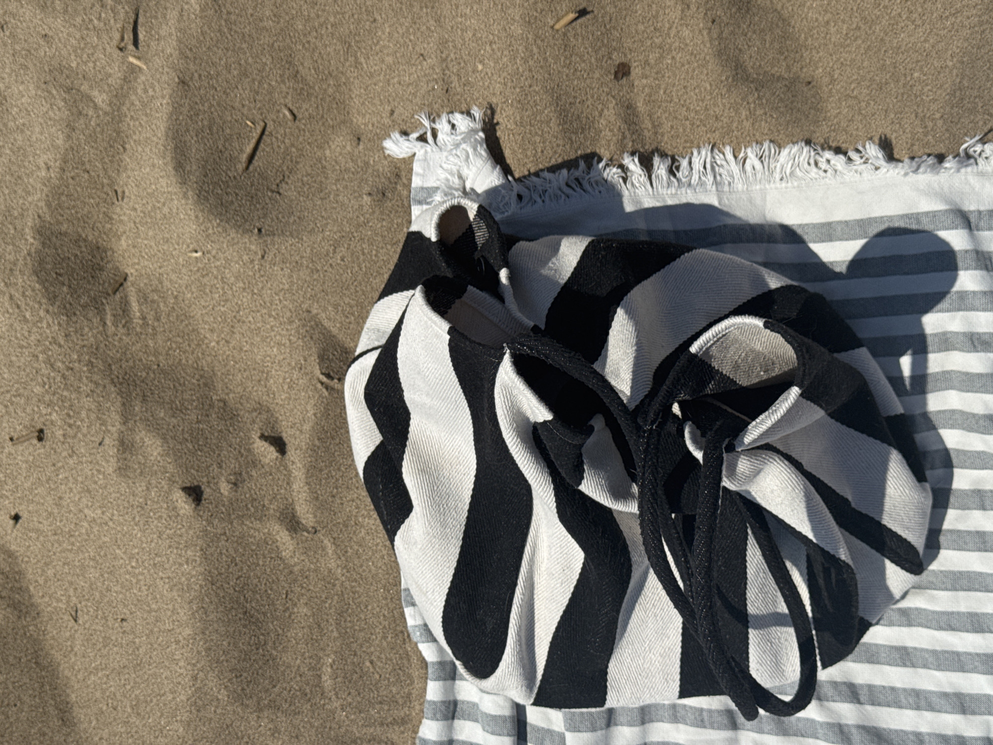 A beach scene shows a black and white striped bag on top of a striped towel on sand. The bag has a drawstring closure. The towel has a fringed edge. The sand is light brown and has some small pieces of debris. The location is unknown; Saratov, Russia cannot be confirmed from this image.