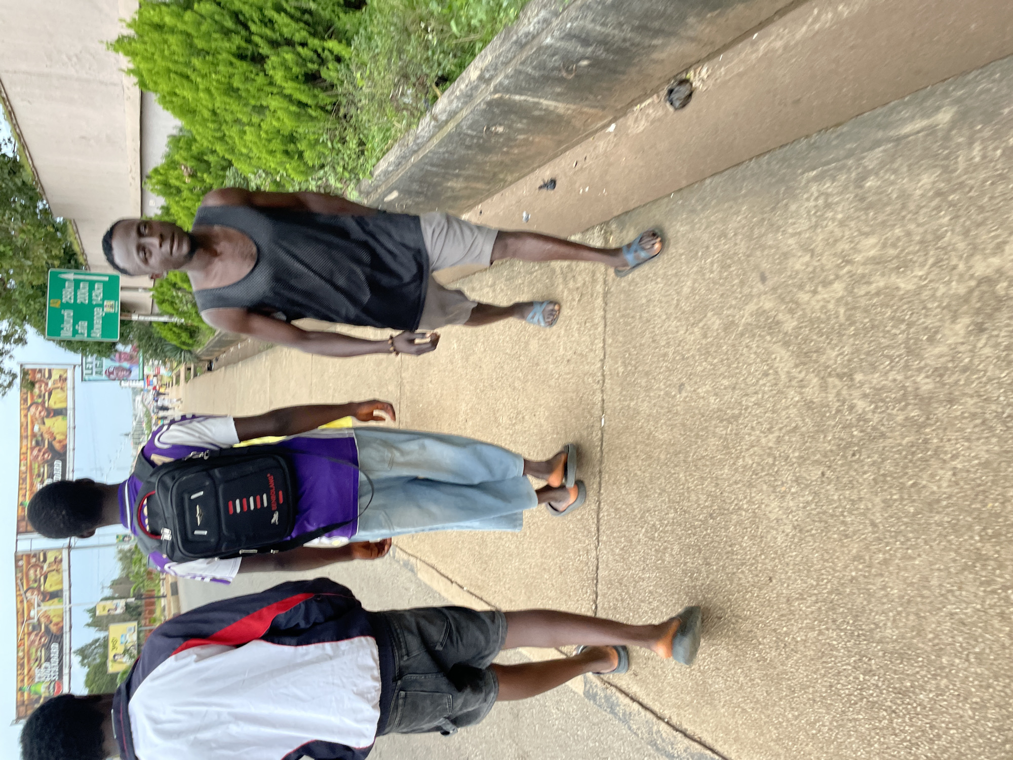 The image captures a street scene in Jos, Nigeria, focusing on three individuals walking along a sidewalk. In the foreground, a person in a black tank top and grey shorts walks towards the camera, their left foot visible in a blue and orange sandal. To their left, a second person, seen from the back, wears a purple t-shirt and light blue denim shorts, also in orange sandals. The third individual, farthest to the left and also seen from the back, is in a white, red, and blue jacket and dark shorts. They are carrying a black backpack with red and white accents.
The environment includes a concrete sidewalk with visible texture and a concrete drain or barrier to the right. The background features lush green bushes lining the barrier, and further back, buildings and large billboards are visible. One billboard displays advertisements, and a green street sign with white text indicating distances to various locations, including "Makurdi 258km" and "Lafia 200km," is prominent. The sky is overcast, suggesting a cloudy day. The overall impression is that of a typical urban street scene in Jos, Nigeria, with people going about their daily activities.