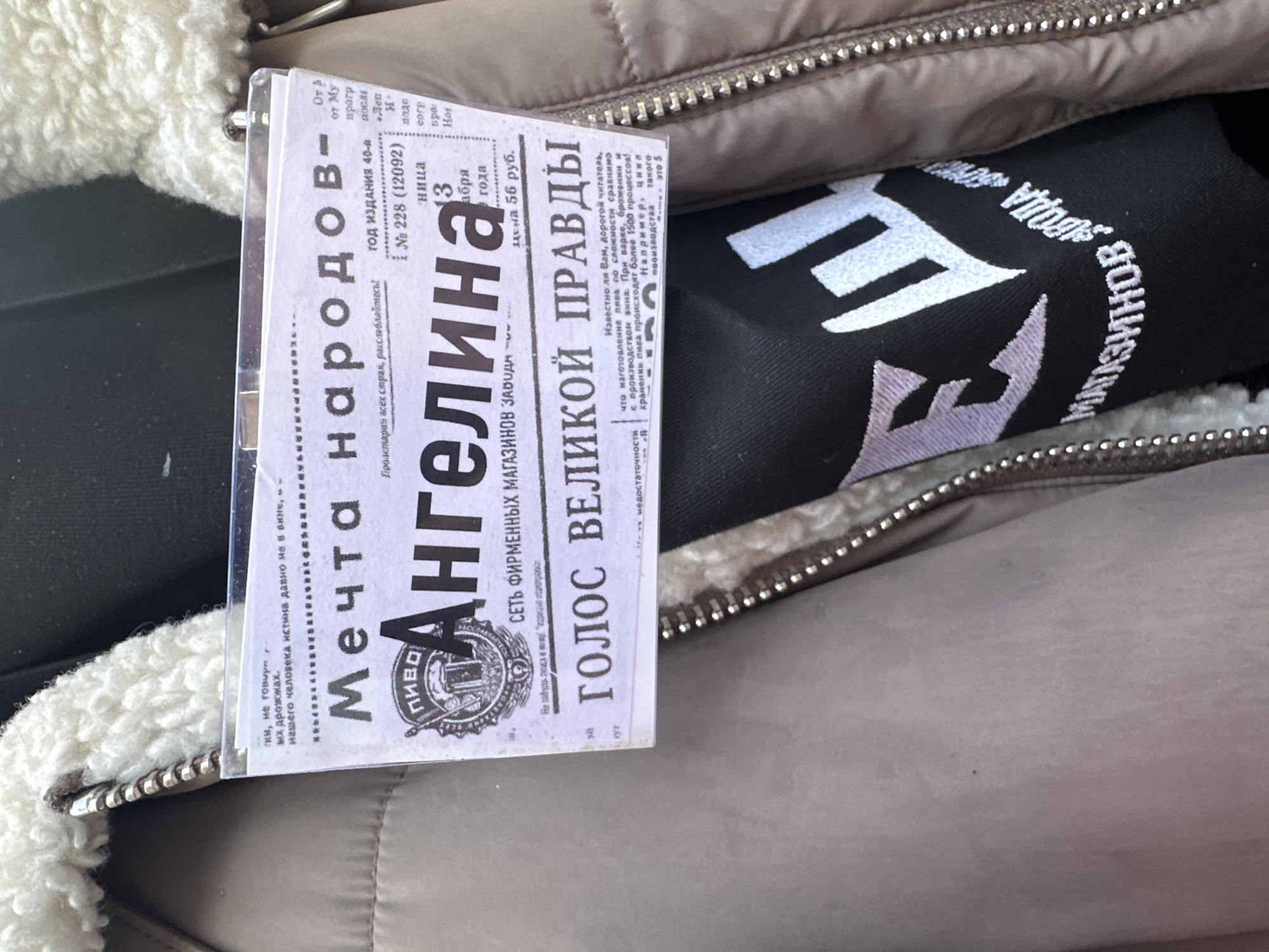 The image shows a close-up shot taken inside a bag or purse. The bag appears to be made of a light brown, quilted material, possibly a jacket or coat, with a visible zipper. The inside lining has a fuzzy, off-white material. Inside the bag, there is a black item with white embroidered lettering. Overlapping this item is a small, laminated newspaper clipping or a print resembling a newspaper advertisement. The headline includes the words "Мечта народов" (Dream of Nations), "Ангелина," and "Голос Великой правды" (Voice of Great Truth). The clipping has text in Russian, indicating the location is likely in Russia. Based on the Cyrillic text and the reference to a "network of branded shops of the plant," this could be a modern imitation designed to look like an old Soviet-era advertisement. Without further context, the exact location in Moscow is impossible to determine, but the use of Russian and the newspaper text suggests that the image was taken there.