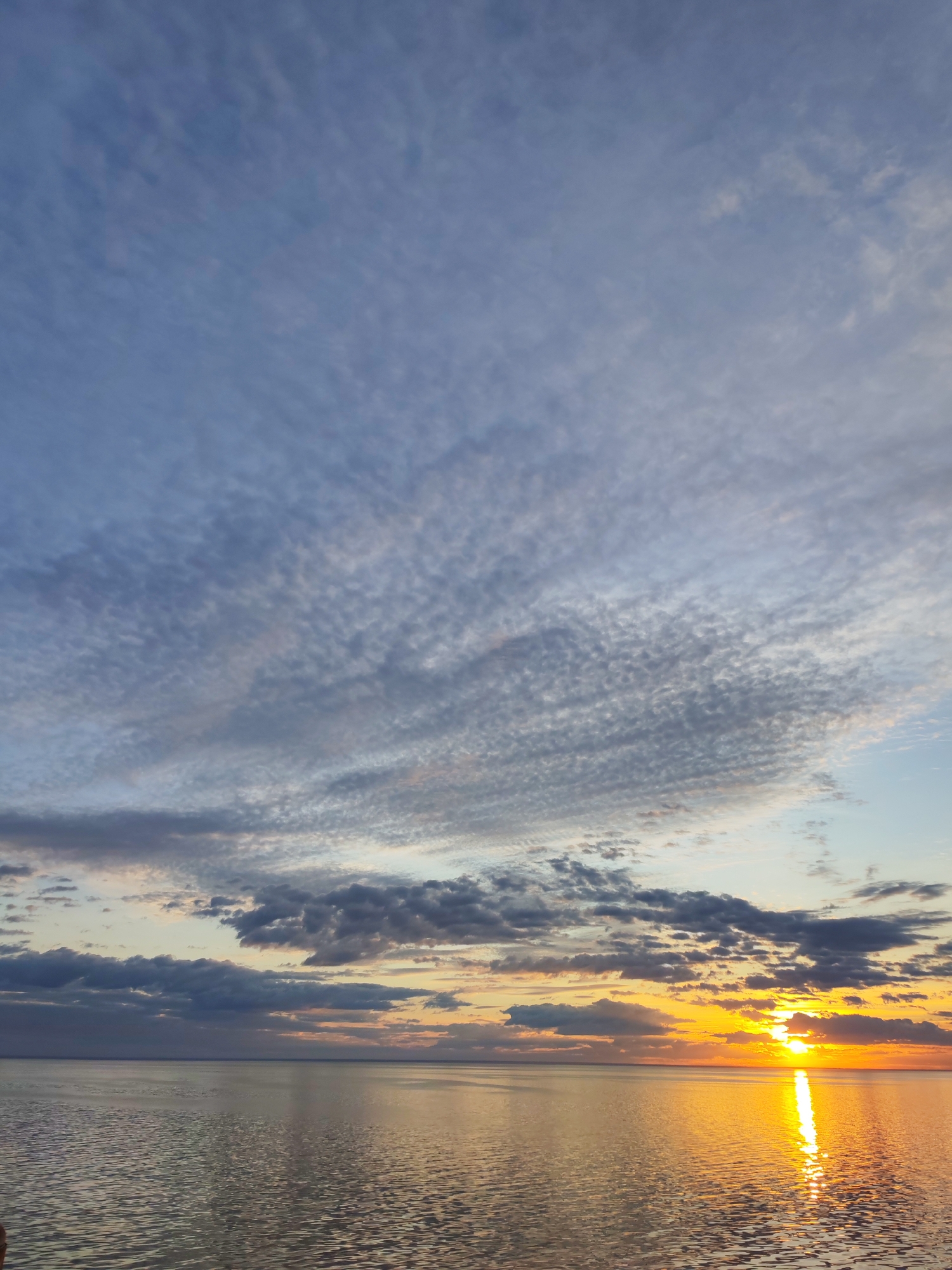 The image shows a sunset over a body of water, likely a lake or sea, in Bol'shie Mkhi, Russia. The sun is low on the horizon, casting a golden reflection on the water's surface. The sky is filled with various types of clouds, including high, thin clouds that create a textured pattern across the upper portion of the sky, and thicker, darker clouds closer to the horizon. The water appears relatively calm, with gentle ripples reflecting the light. There are no discernible people or objects in the immediate foreground.