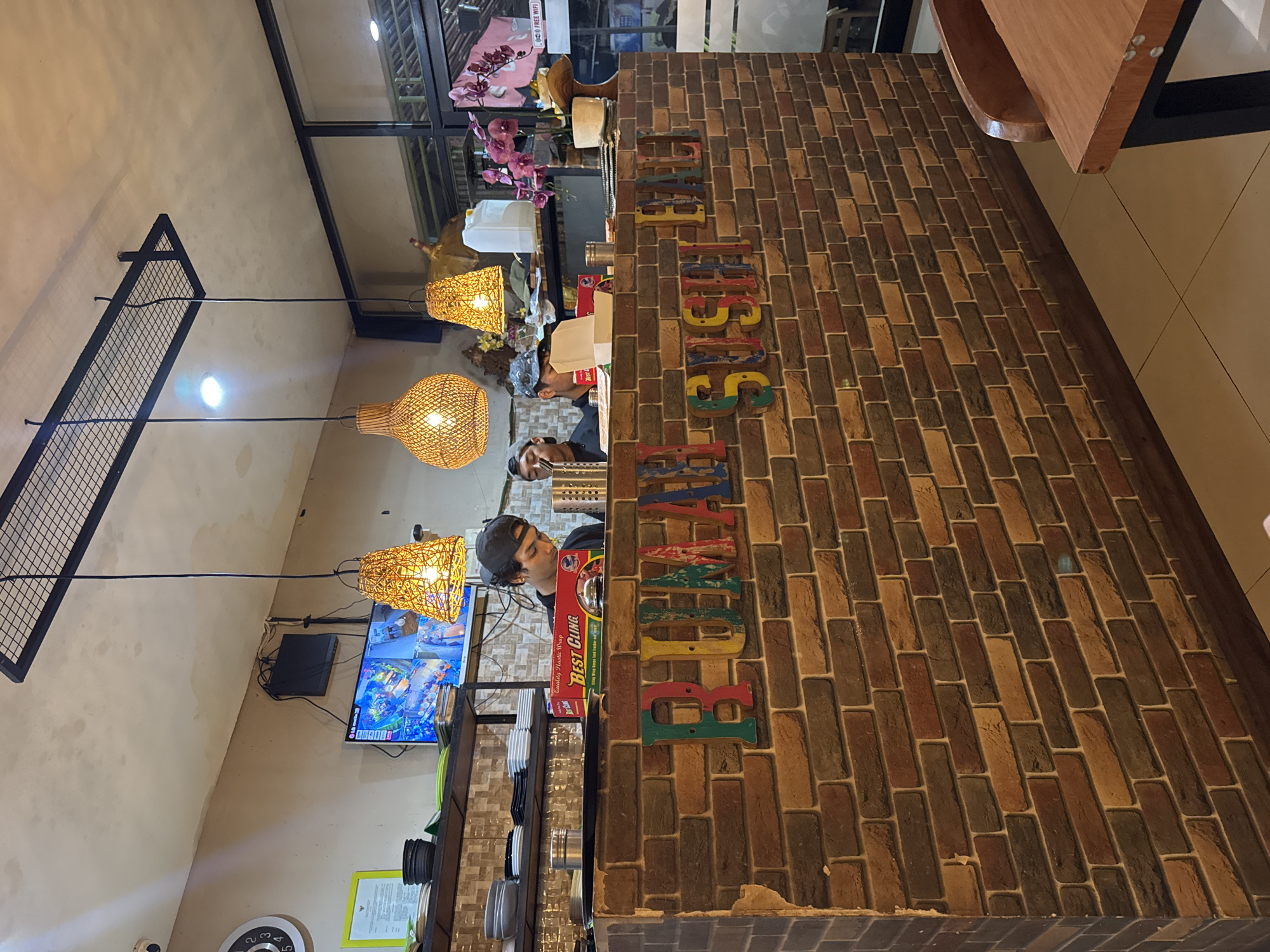 The image depicts the interior of a sushi restaurant in Kuta Selatan, Indonesia. The foreground is dominated by a brick counter with colorful, distressed wooden letters spelling out "RUMAH SUSHI BALI." Behind the counter, two individuals, likely staff, are visible. The person on the left, wearing a black t-shirt and a baseball cap, is looking down and appears to be working. Another person, partially obscured, stands to their right.
Above the counter, shelves are stocked with plates and bowls. A television screen displays multiple camera feeds, suggesting a security or surveillance system. Three woven pendant lamps hang from the ceiling, casting a warm light. The background reveals large windows, with a glimpse of outdoor seating and foliage. In the middle ground, a white container and a bouquet of pink orchids add decorative elements. The overall atmosphere suggests a casual dining establishment.